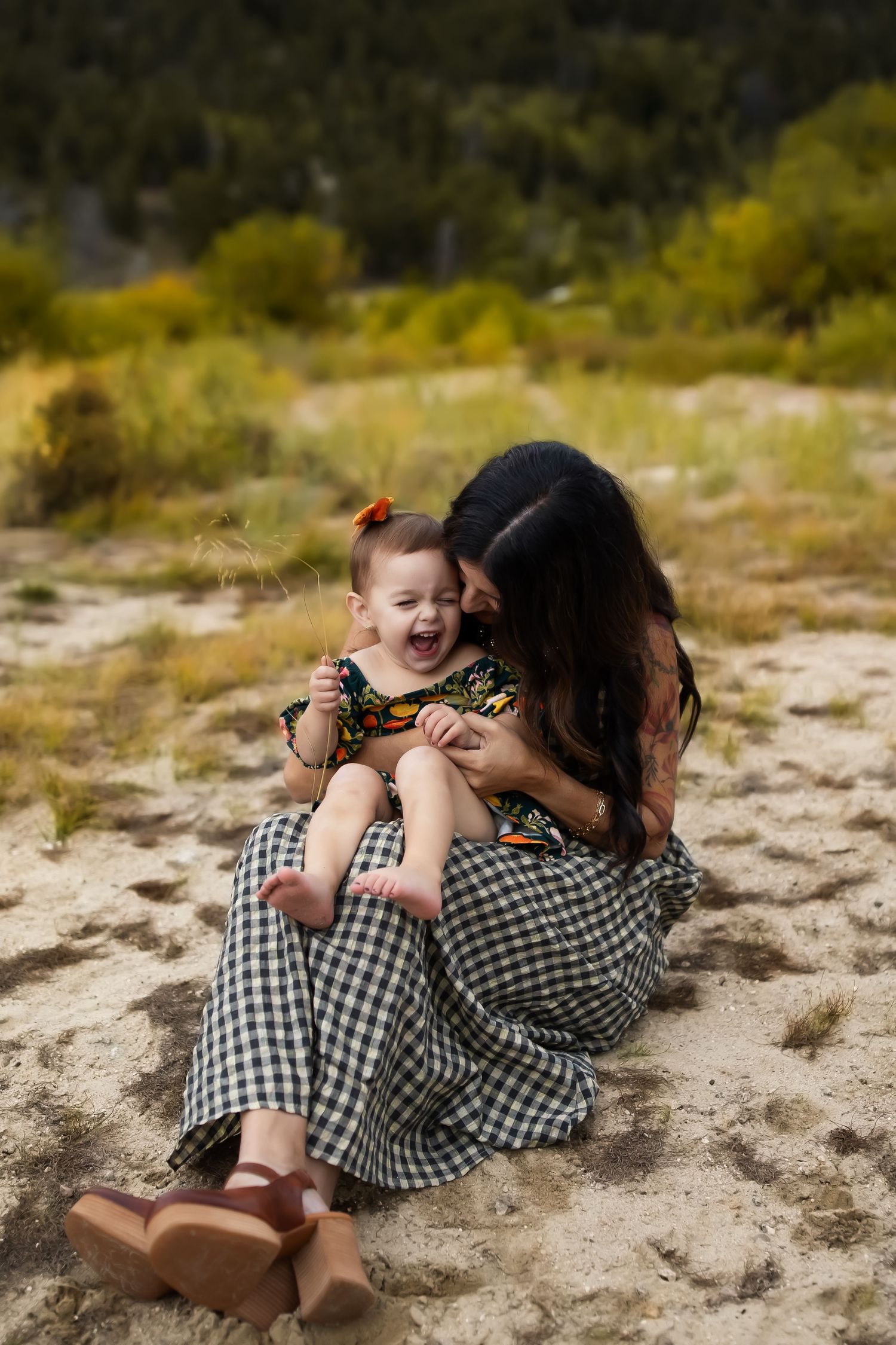 Two people in gingham clothing share a playful moment while lying on sandy ground with mountains in the background.
