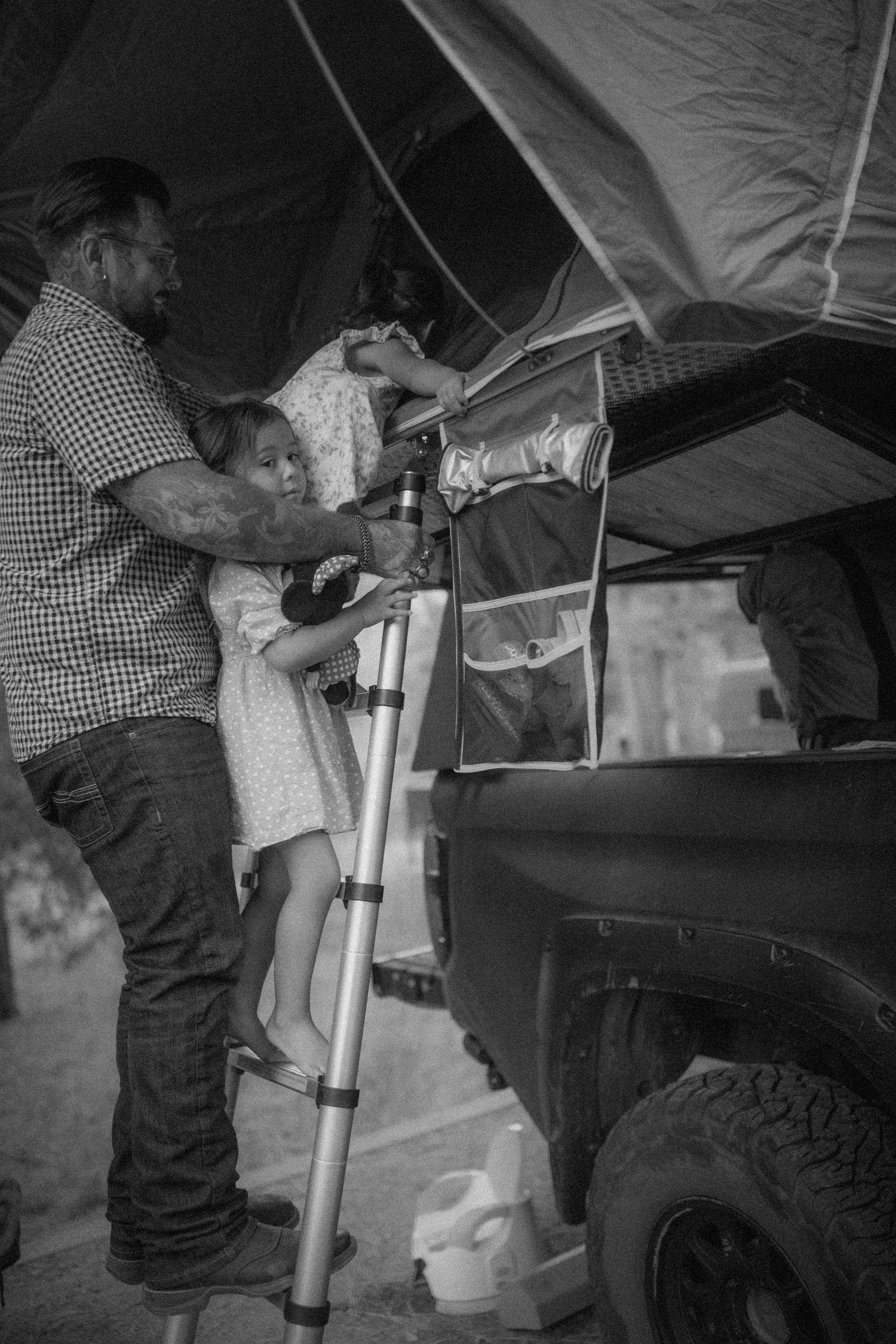 Setting up camping equipment near a vehicle with a rooftop tent in black and white.