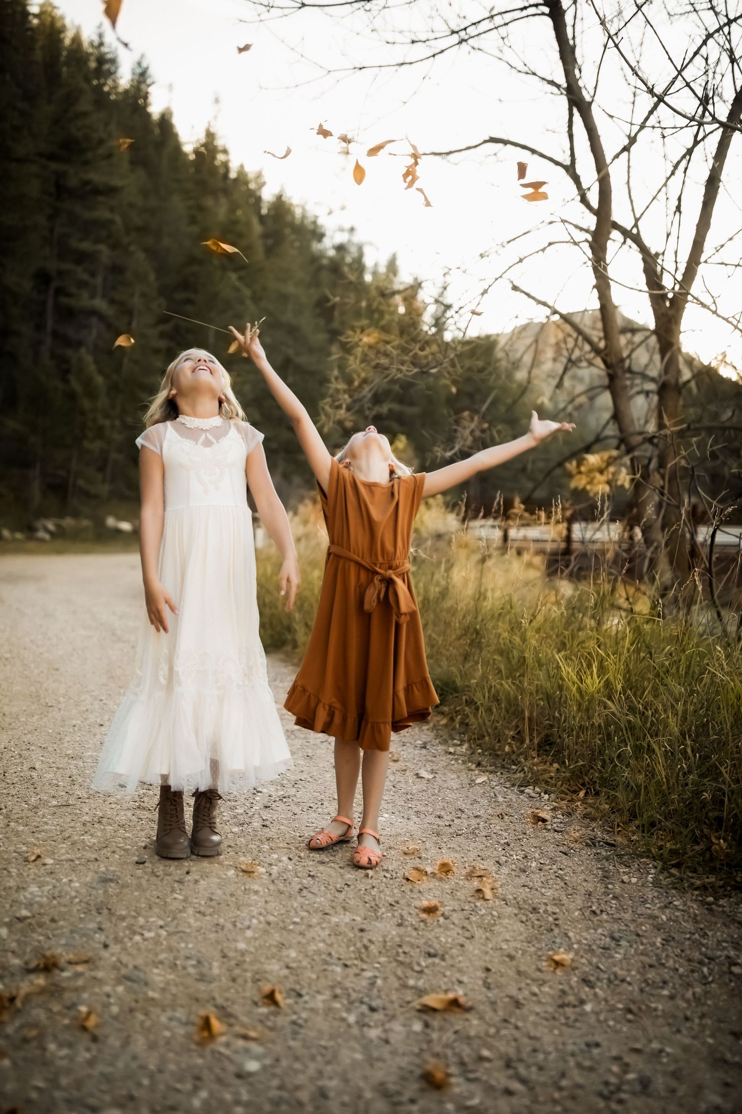 Two figures in white and brown outfits playfully toss autumn leaves on a forest path.