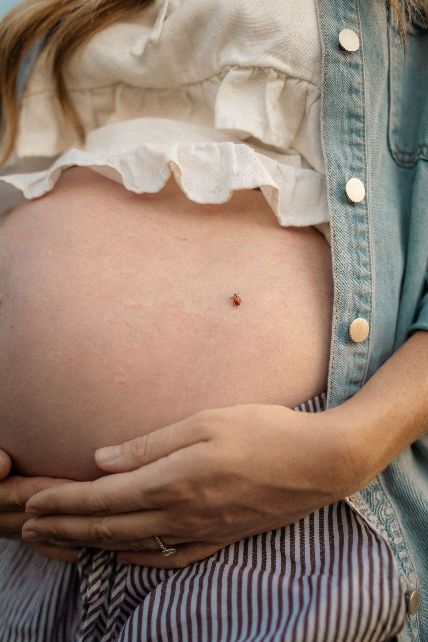 A pregnant woman in denim and white clothing cradles her baby bump while standing in soft natural light.