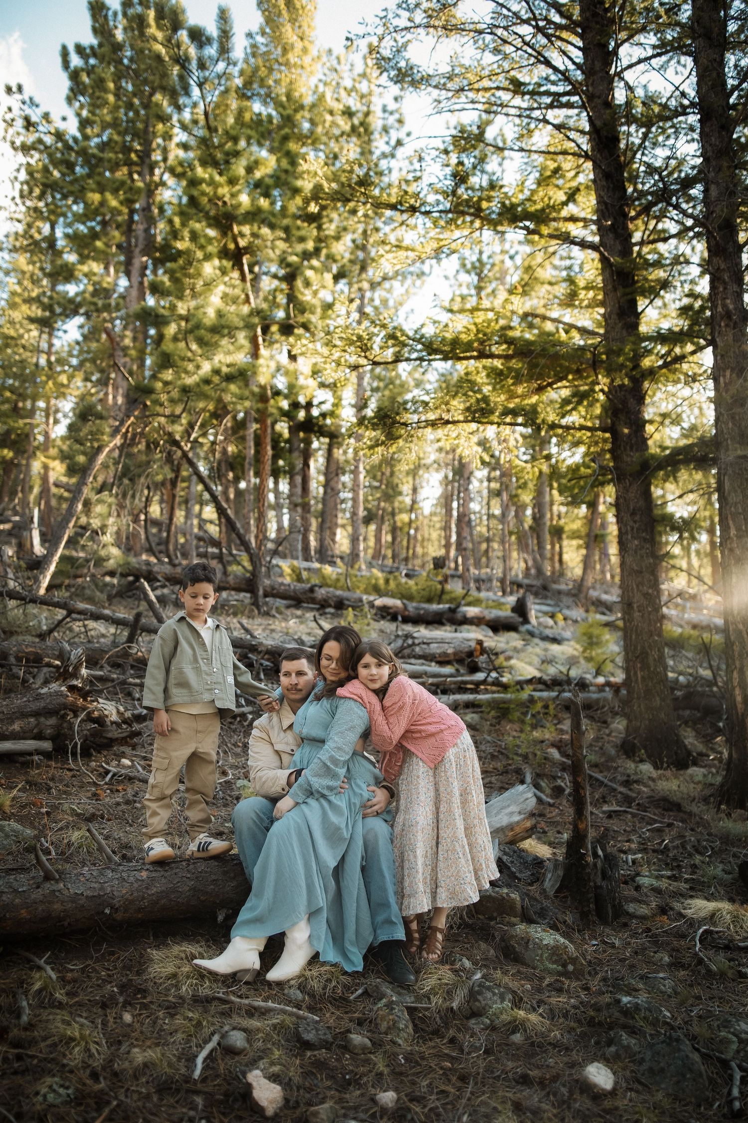 Family of four poses together on a fallen log in a sunlit pine forest, wearing elegant earth-tone outfits.