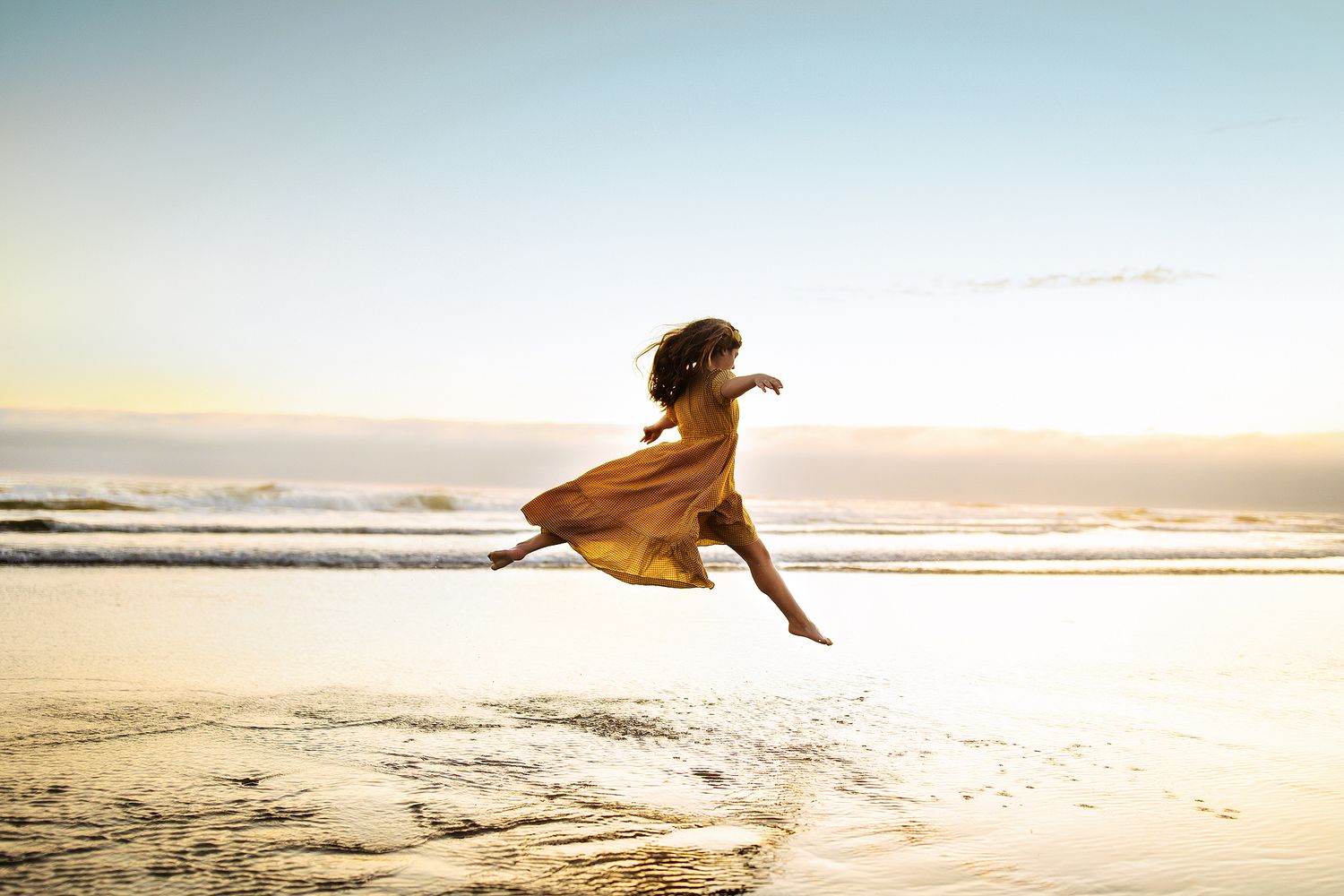 Person in flowing dress runs and jumps along beach at sunset.