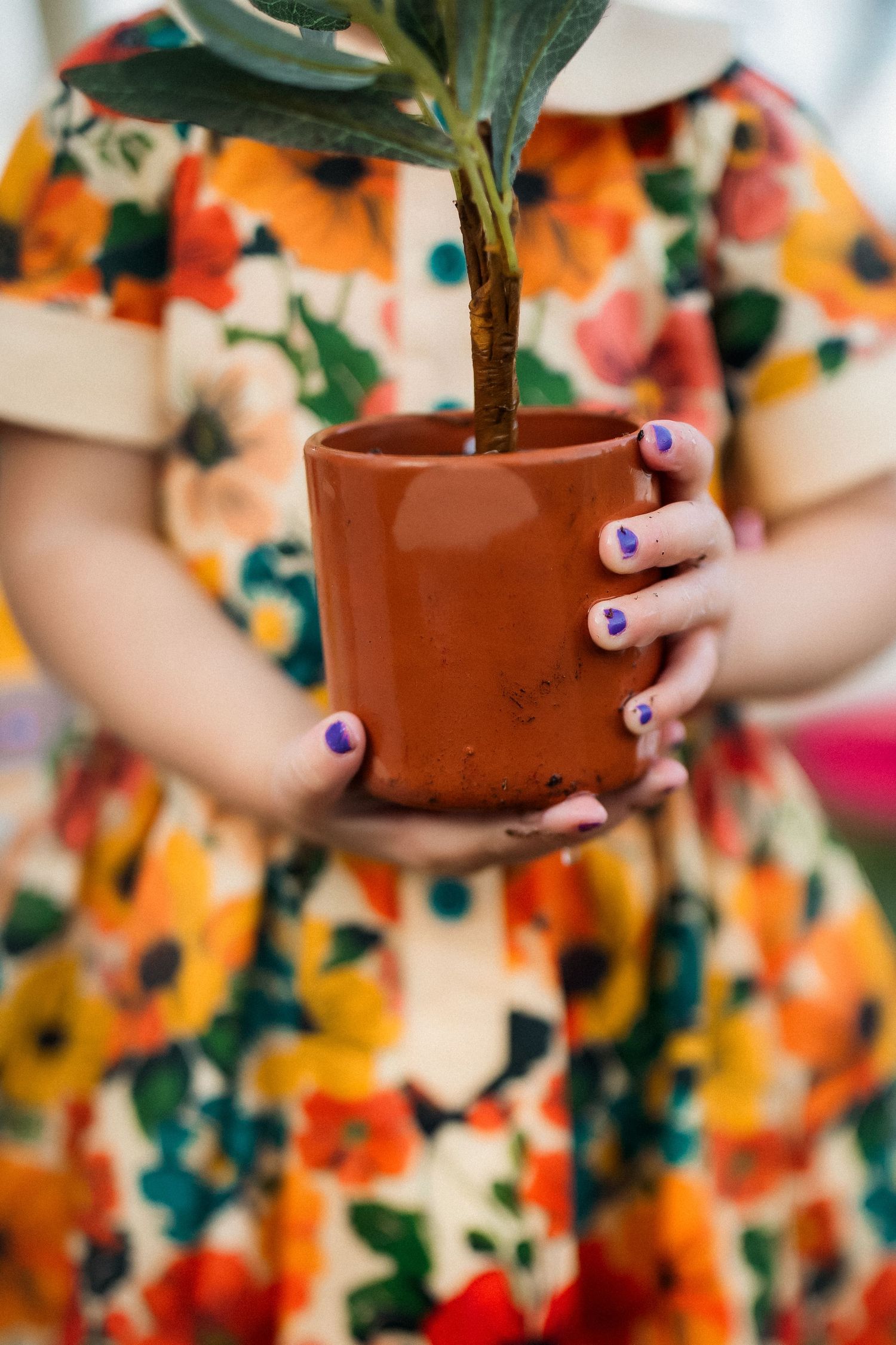 Child in floral dress holding a terracotta plant pot with purple painted nails.