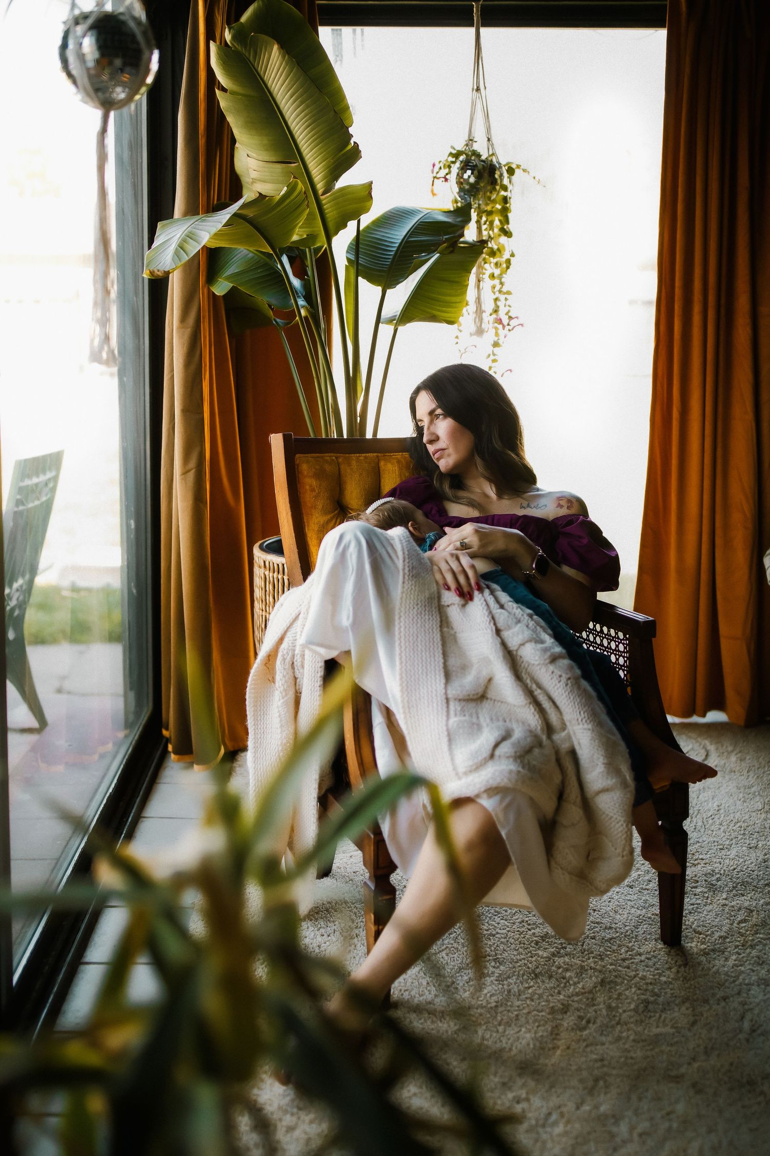 Woman sitting in a wooden chair by a window, wrapped in a blanket, surrounded by tropical plants and orange curtains.