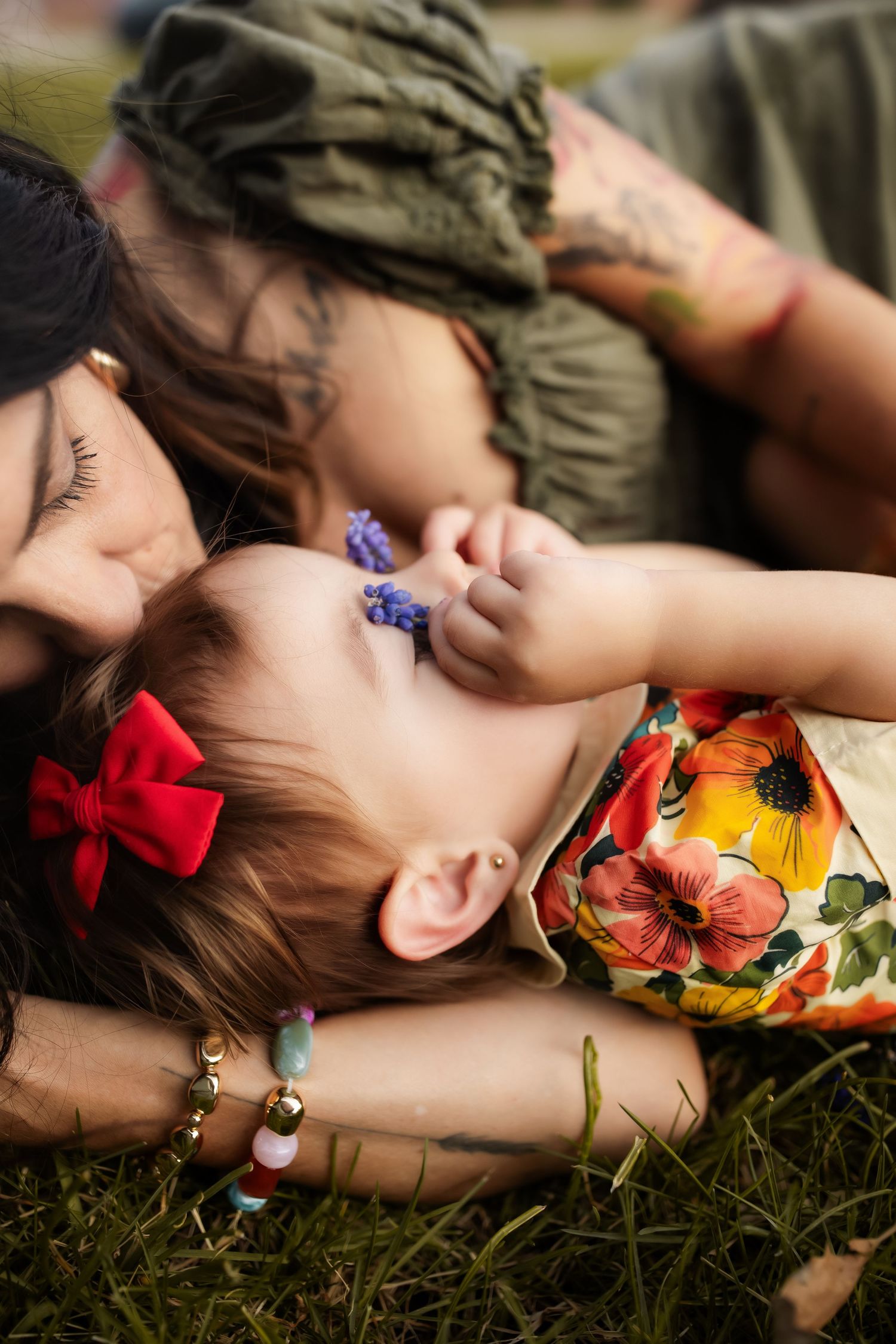 A mother and young child lying on grass, sharing a tender moment, child wearing floral dress with red bow in hair.