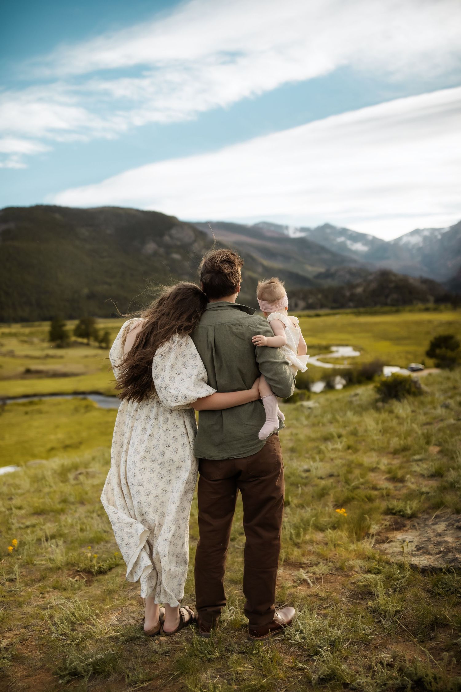 A family of three stands together gazing at a scenic mountain valley, mother in floral dress, father holding baby.