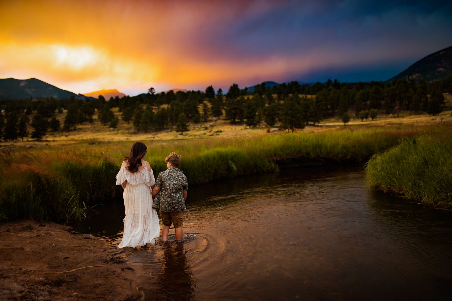 A couple stands in shallow water against a dramatic sunset in the mountains.
