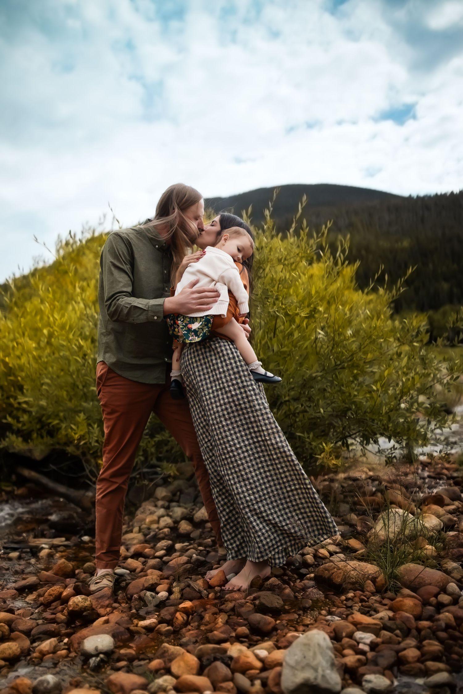 A couple embraces on rocky terrain with yellow wildflowers and mountains in the background under cloudy skies.