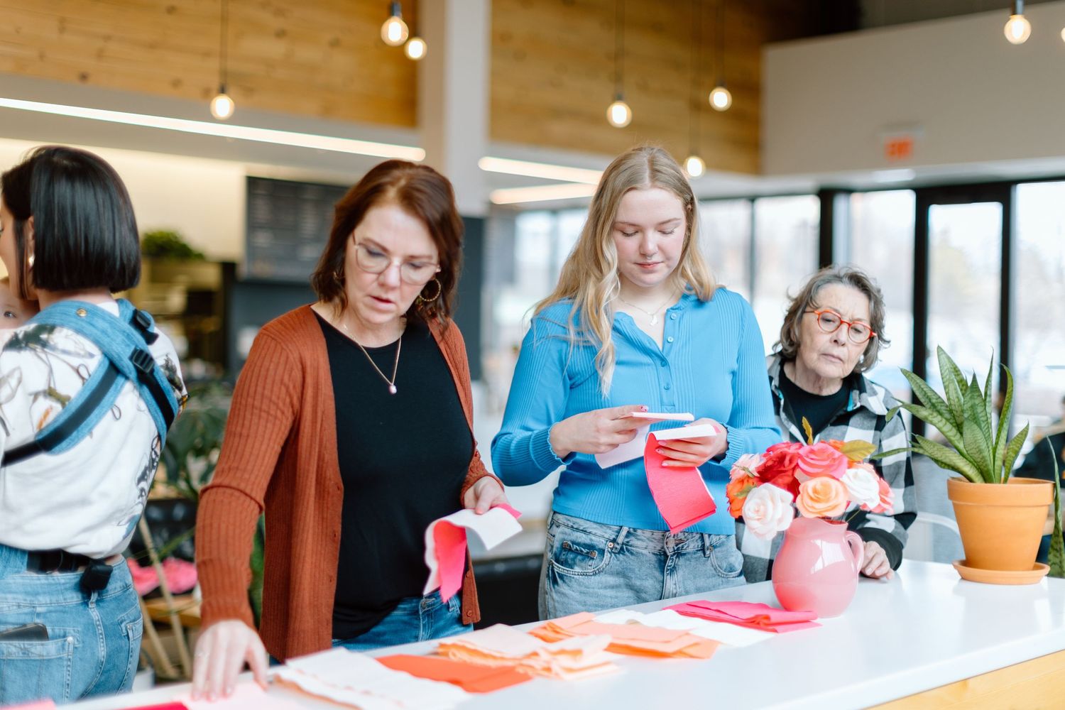Women crafting Valentine's Day decorations at a table with red and pink paper, flowers, and festive supplies indoors.