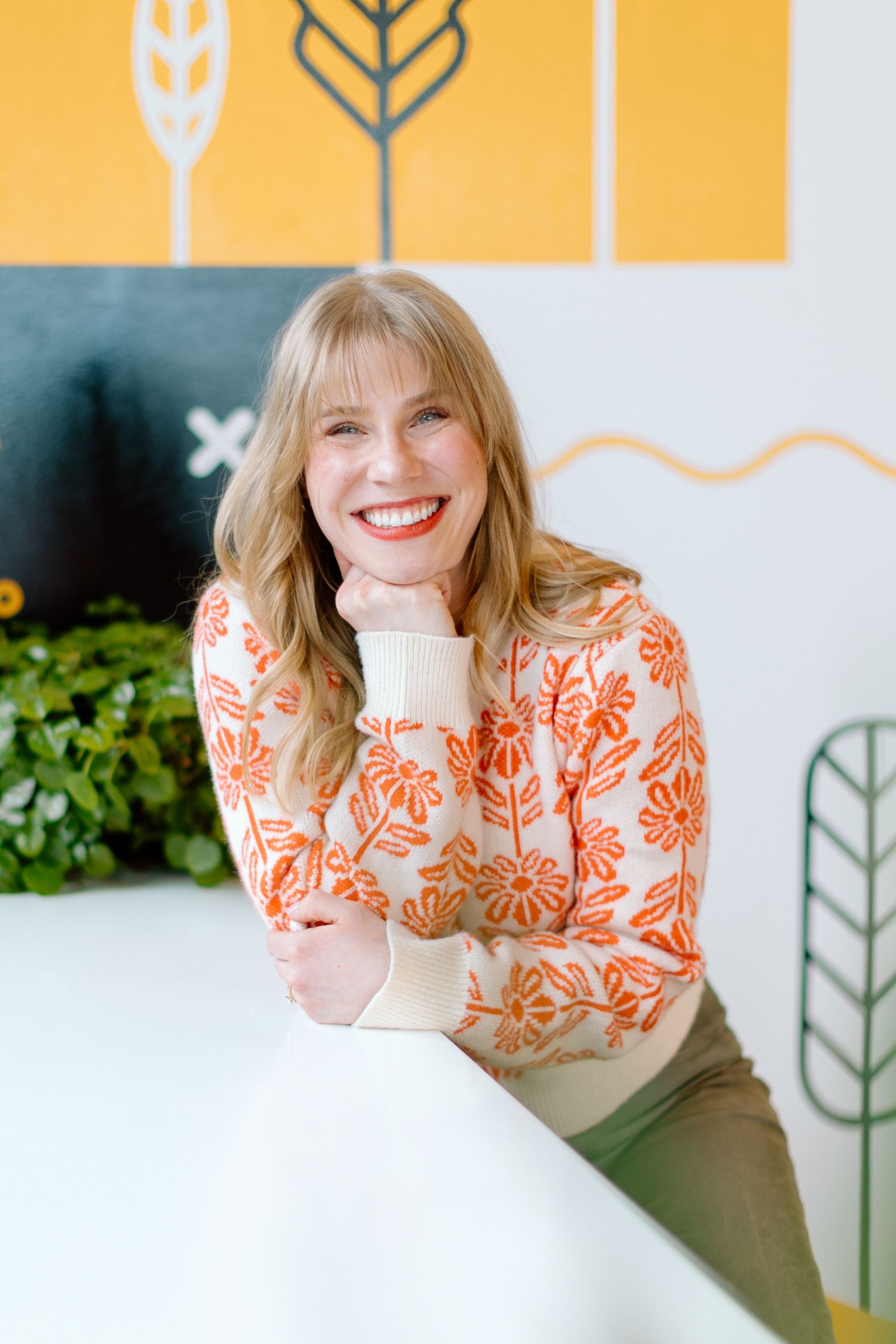 Smiling blonde woman in orange floral sweater leaning on white surface against colorful illustrated wall.