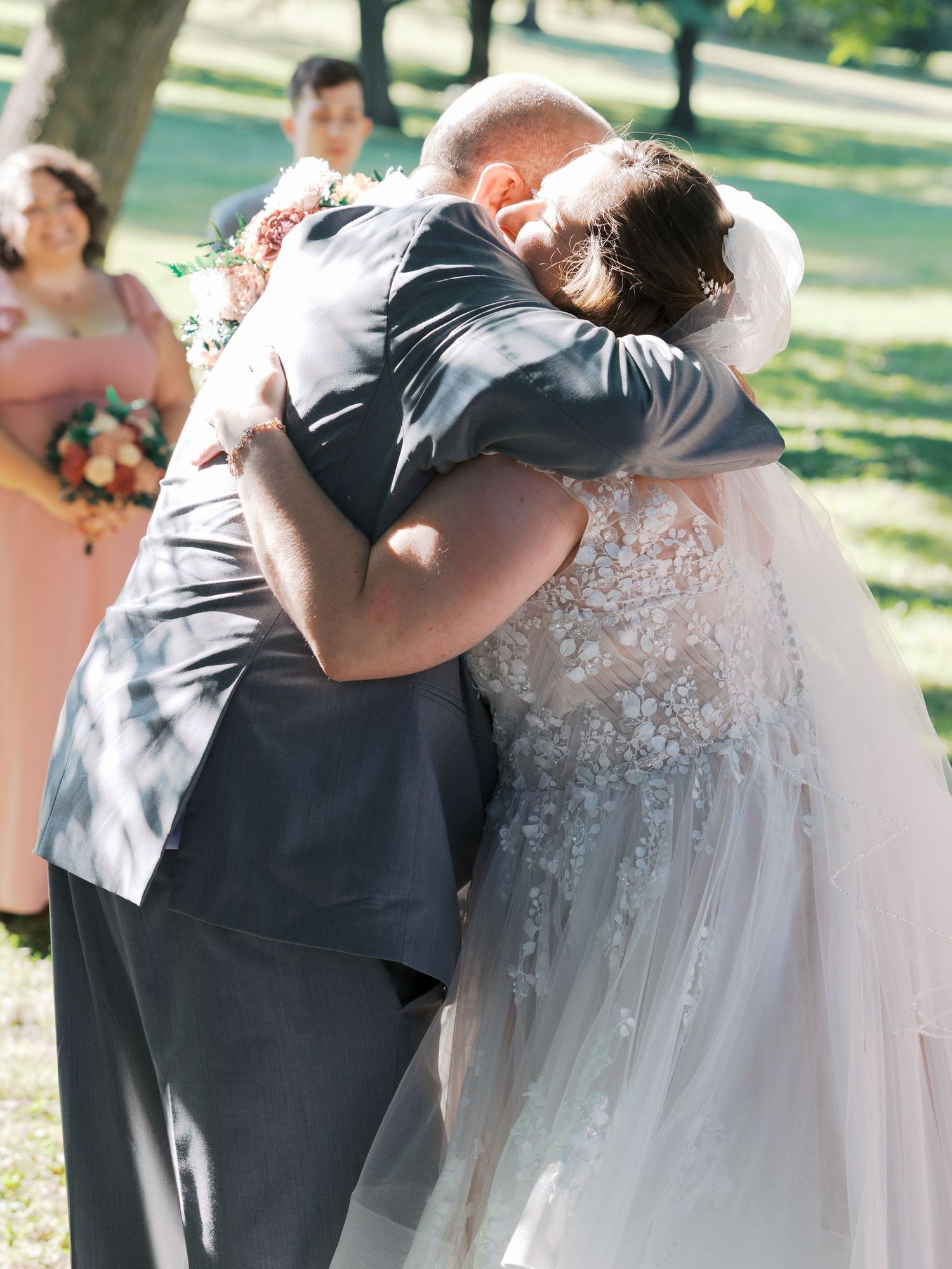 Bride in lace gown shares emotional hug with guest at outdoor wedding ceremony.