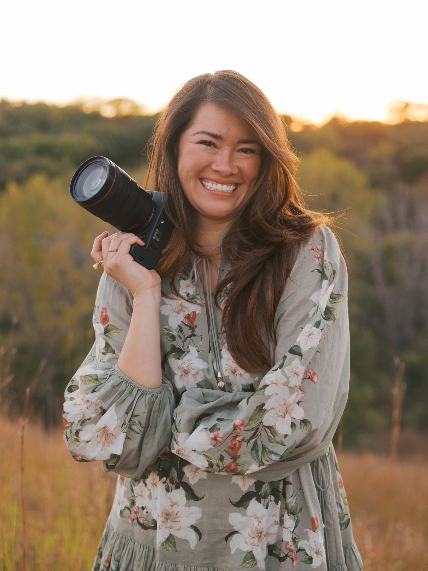 Professional photographer in floral dress poses with camera in golden field at sunset.