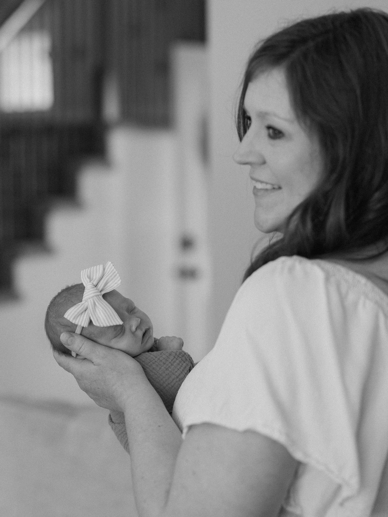 Black and white photo of a smiling woman holding a swaddled newborn baby with a bow headband indoors.