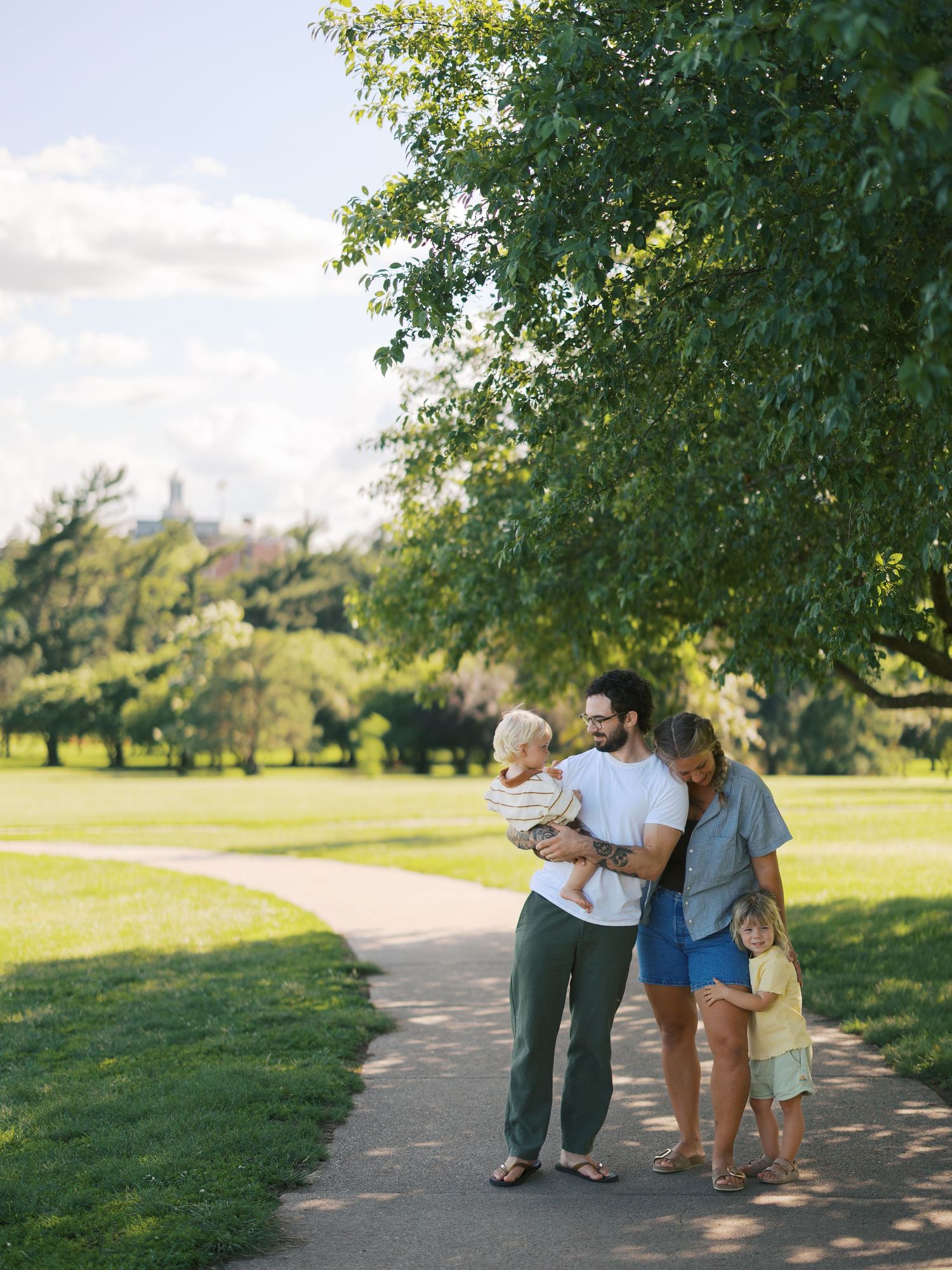 Family of four enjoying a sunny walk on a park path surrounded by lush green trees and open lawns.