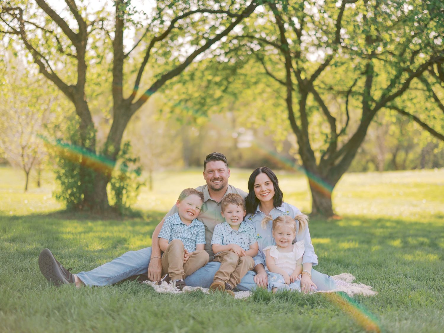 Happy family of five posing outdoors on green grass surrounded by sunlit trees during a spring photo session.