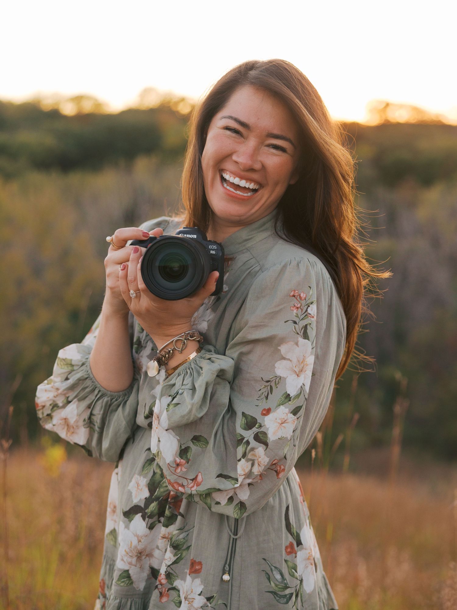 Smiling woman in floral dress holding a DSLR camera outdoors in a golden sunlit field.