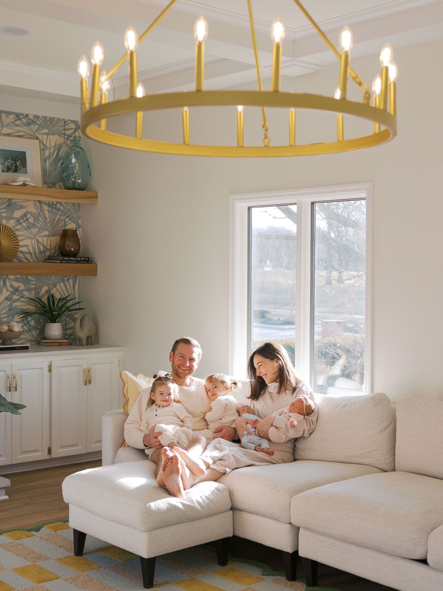 Family of five in matching outfits sitting on a white sectional sofa beneath a gold ring chandelier in a bright living room.