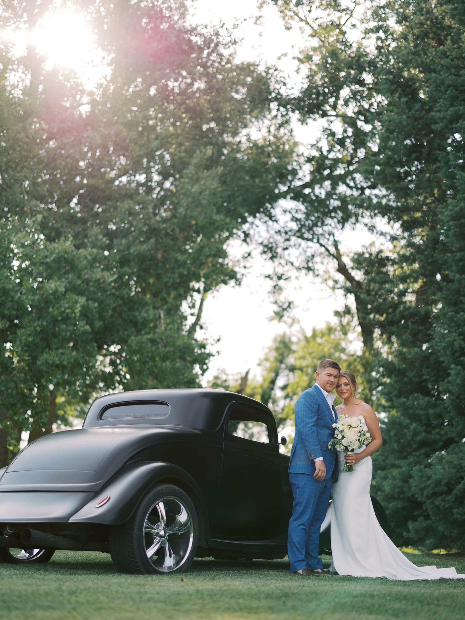 Bride and groom posing beside a vintage black hot rod car surrounded by lush green trees on their wedding day.