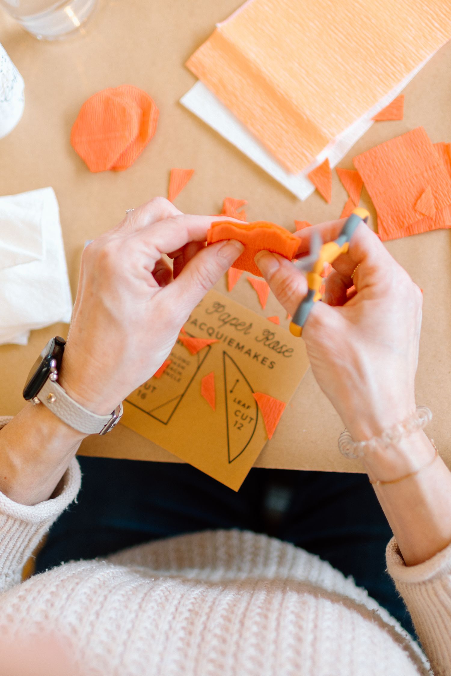 Hands crafting orange crepe paper flowers using a paper flower kit on a wooden table.