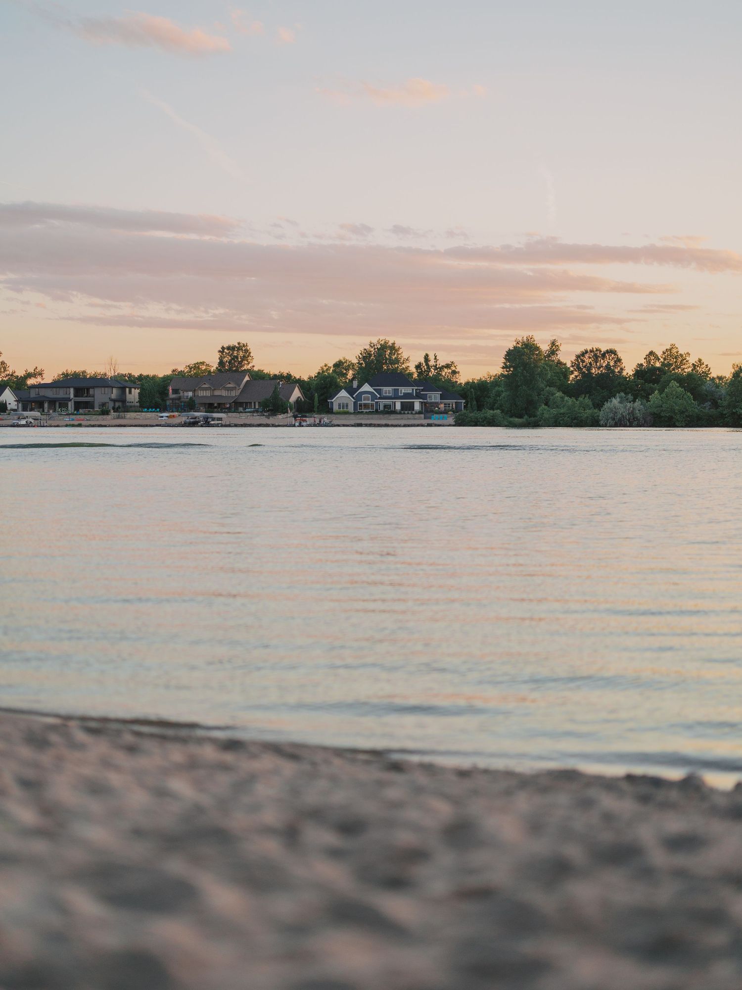 Sandy beach shoreline at golden hour sunset with calm water reflecting pink sky and houses with trees in the background.