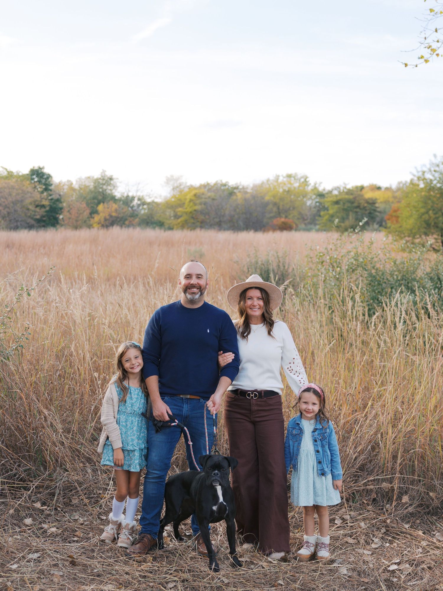 Family of four with black dog posing in golden field during autumn, wearing casual fall outfits.