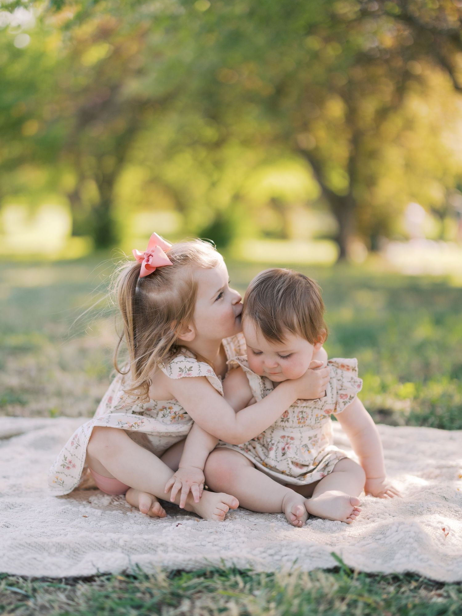 Two young sisters share a sweet kiss on a blanket outdoors, surrounded by lush green trees in soft natural light.