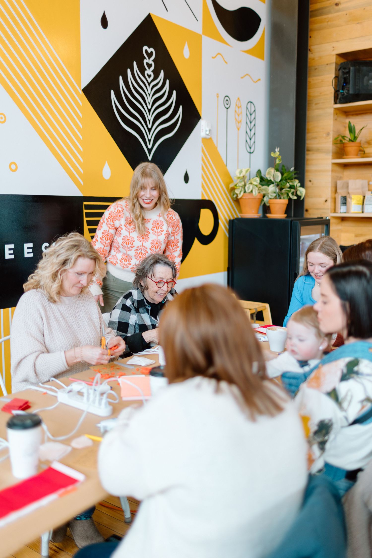 Women gathered around a table crafting in a colorful cafe with yellow geometric wall art and wooden decor.