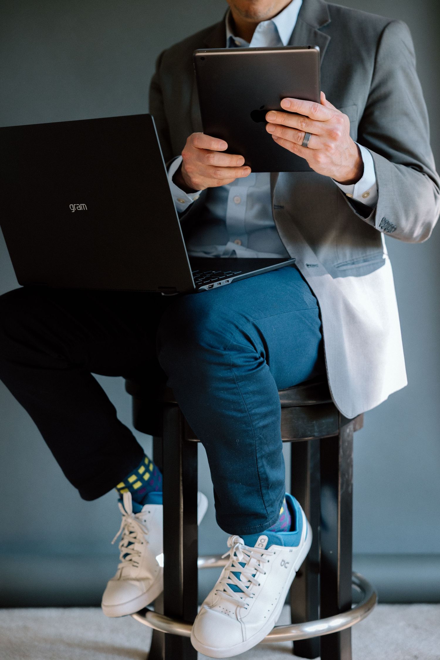 Man in gray blazer and blue pants sitting on stool using laptop and tablet, wearing white sneakers and colorful socks.
