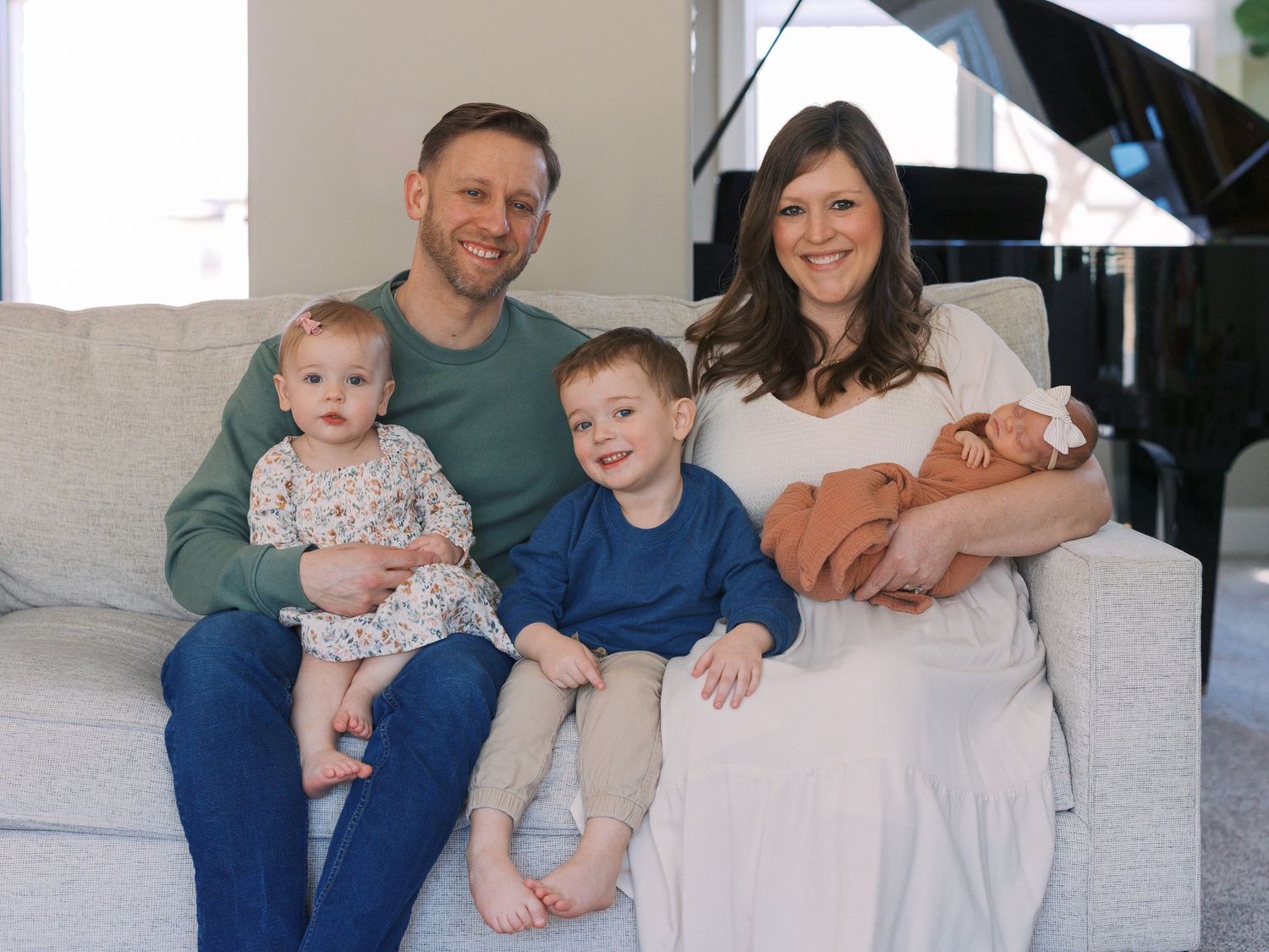 Happy family of five posing on a couch, parents with two toddlers and a newborn baby in a bright living room.