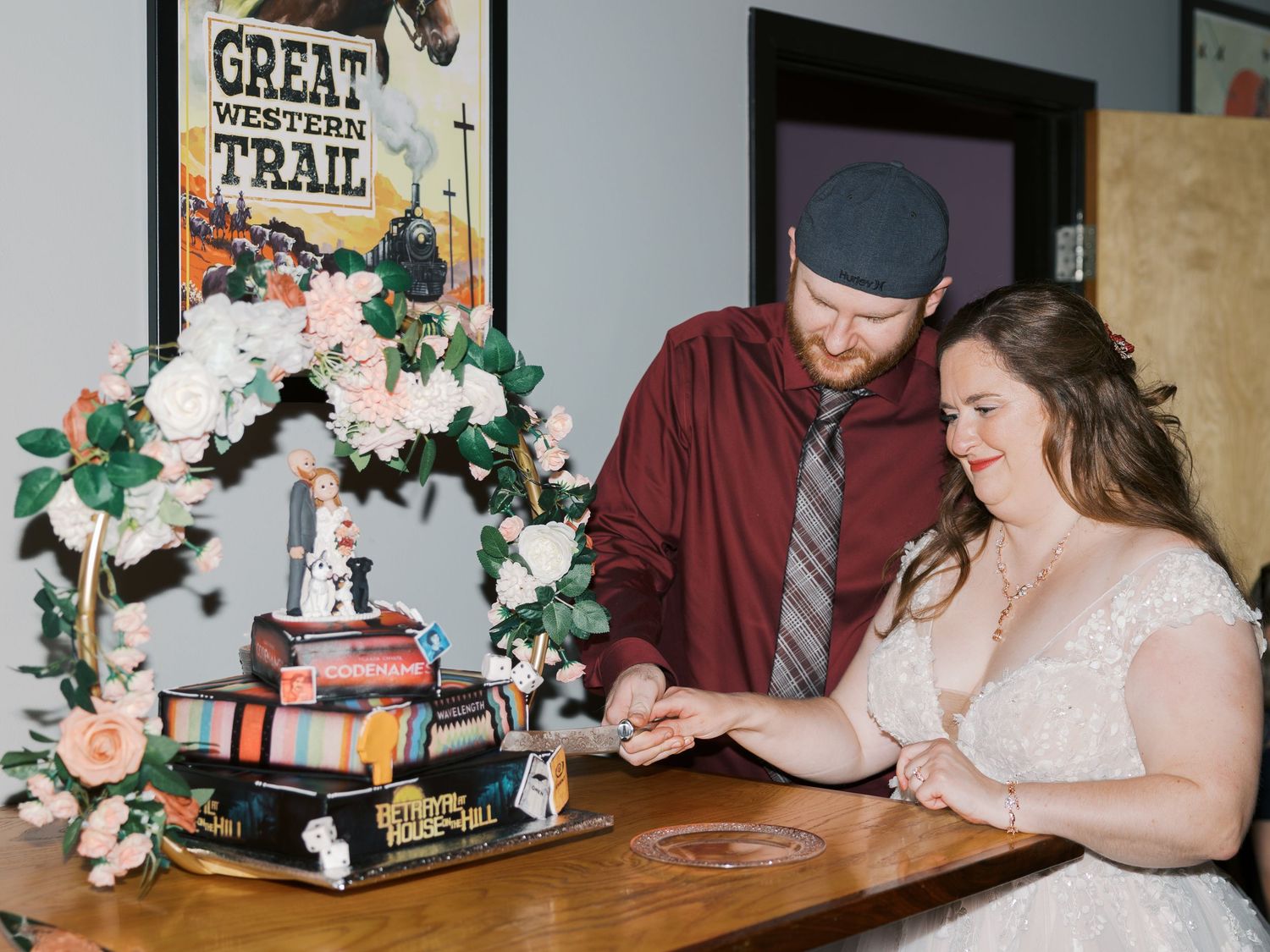 Bride and groom cutting a book-themed wedding cake decorated with floral arch near a Great Western Trail poster.