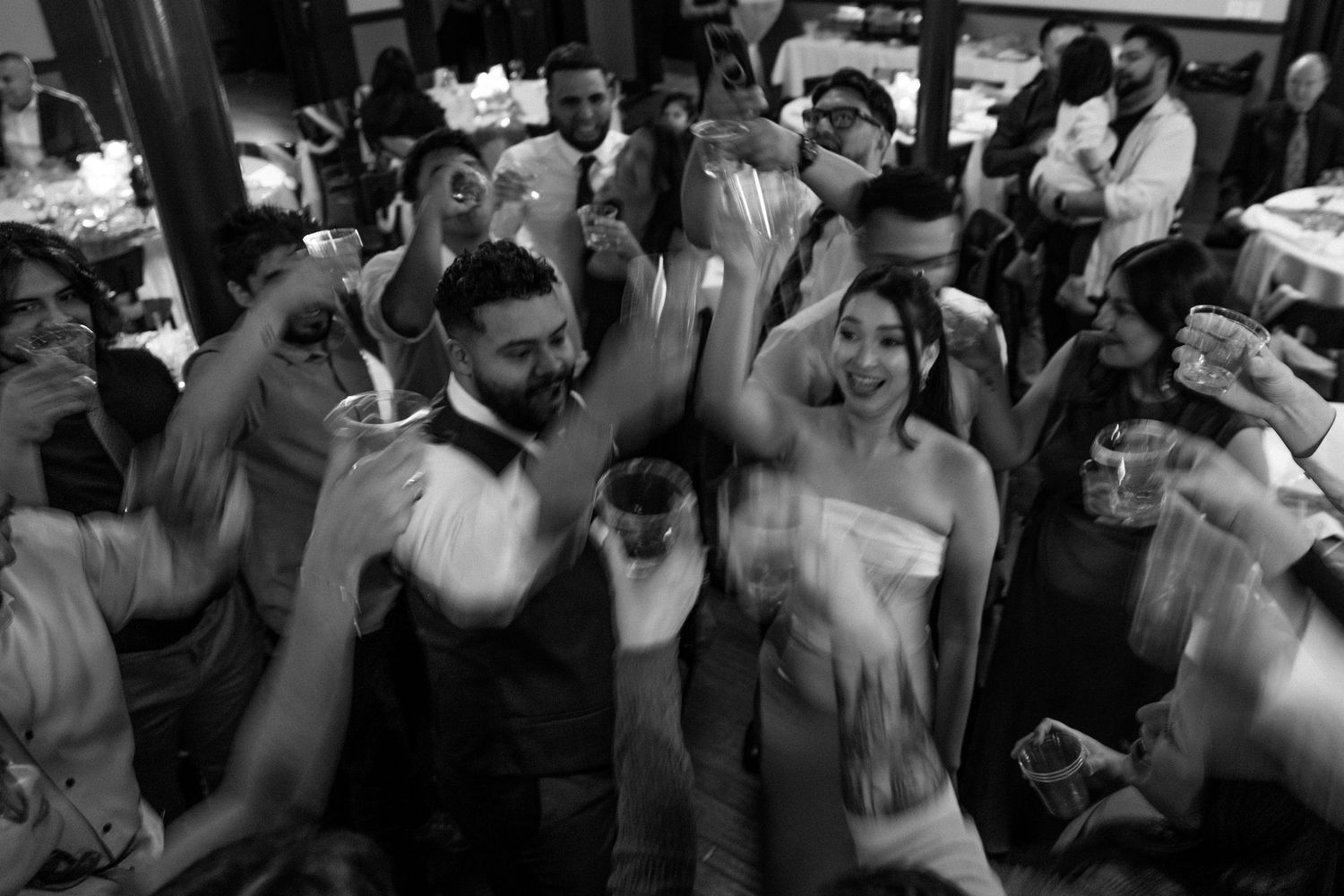 Black and white photo of bride and groom dancing joyfully with guests at a lively wedding reception.
