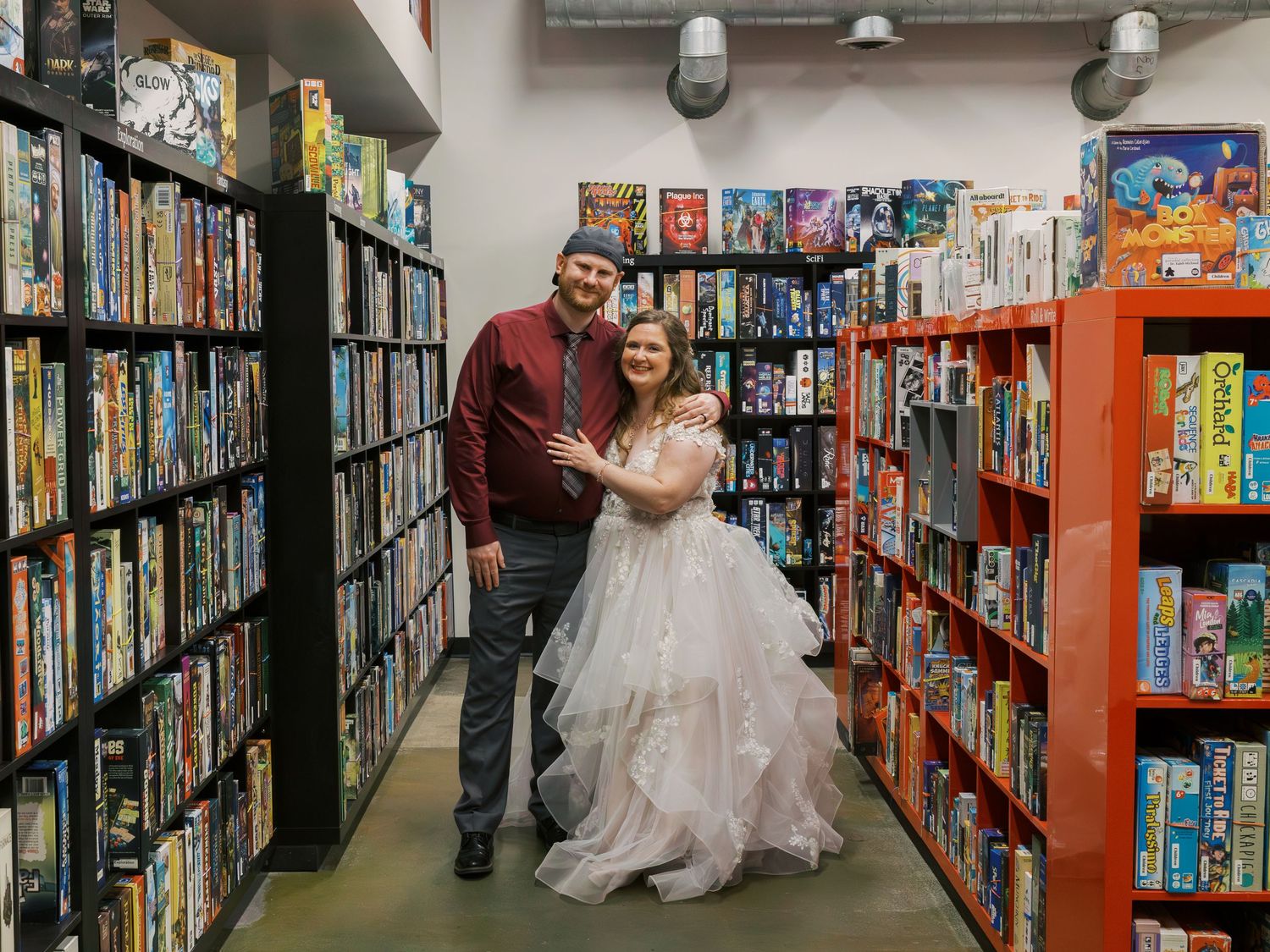 Couple in wedding attire posing in board game store aisle surrounded by shelves of games.