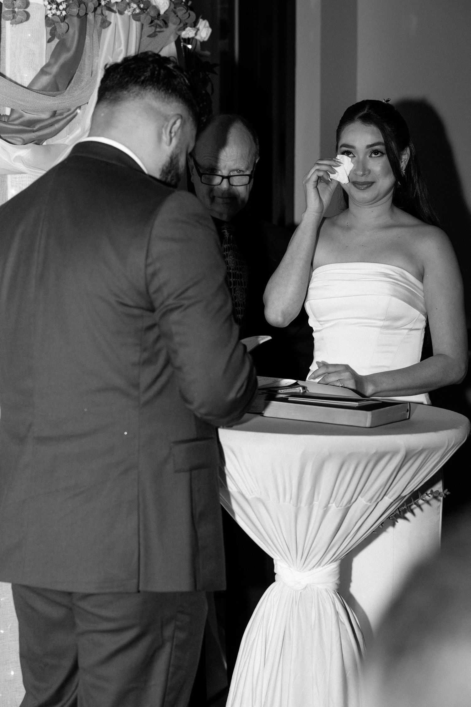 Black and white photo of a bride wiping tears during wedding ceremony while groom signs documents at draped table.