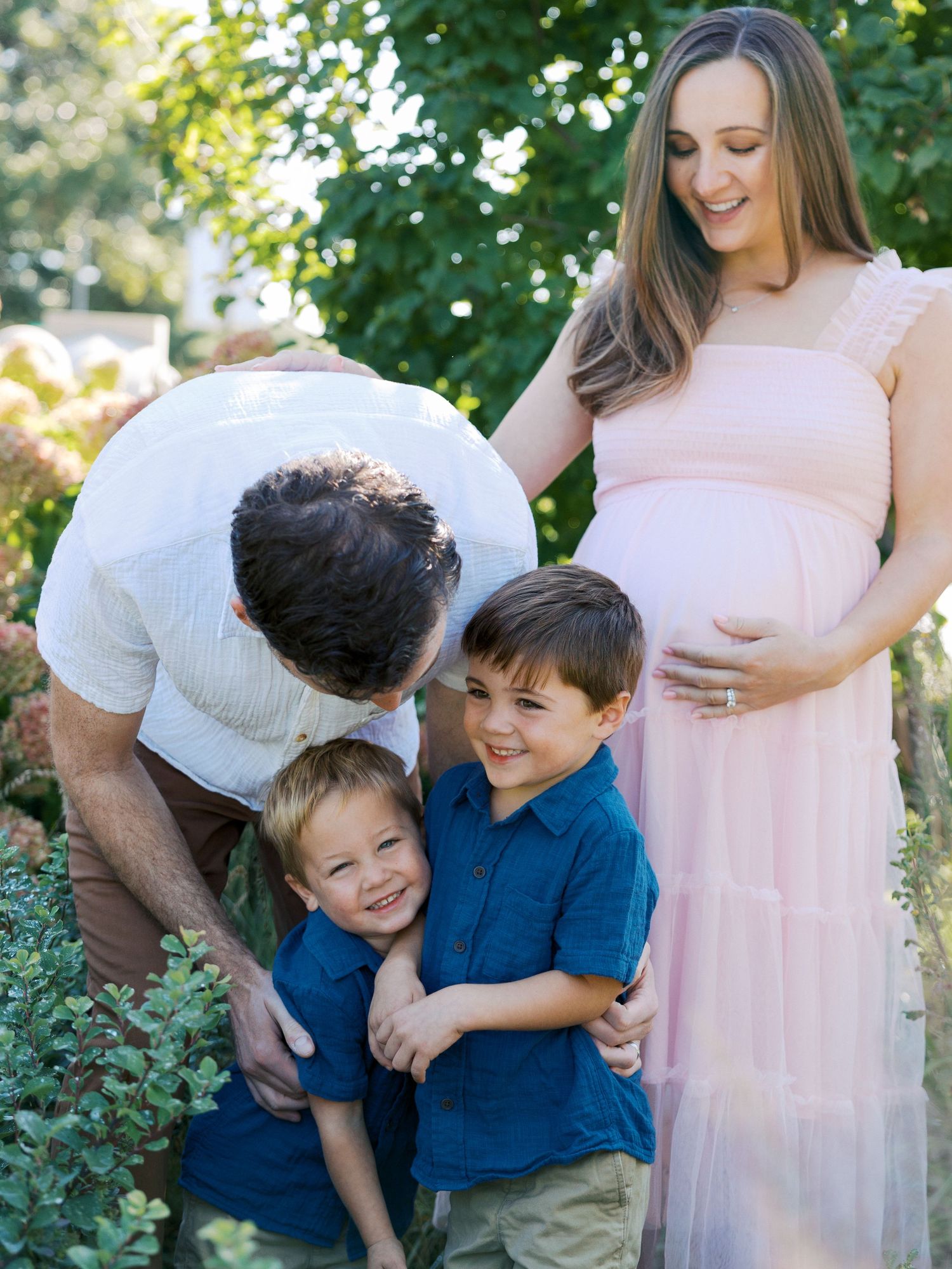 Pregnant woman in pink dress with family, husband hugging two young boys in blue shirts outdoors.