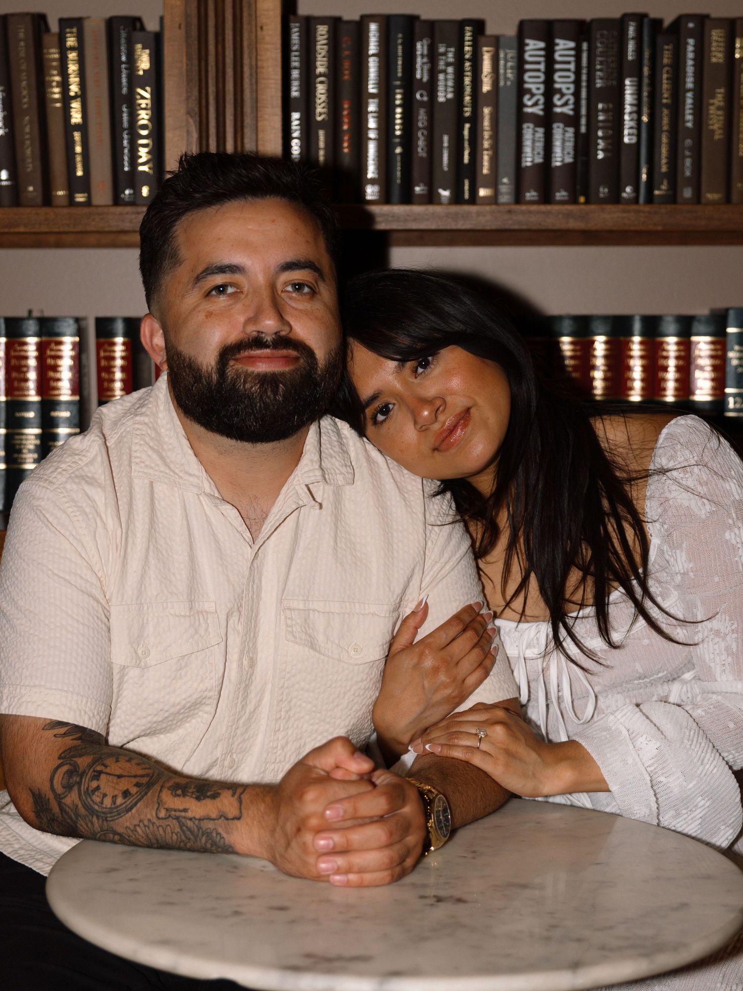Couple posing together at a marble table in front of a bookshelf, man with beard and tattoos, woman in white top.