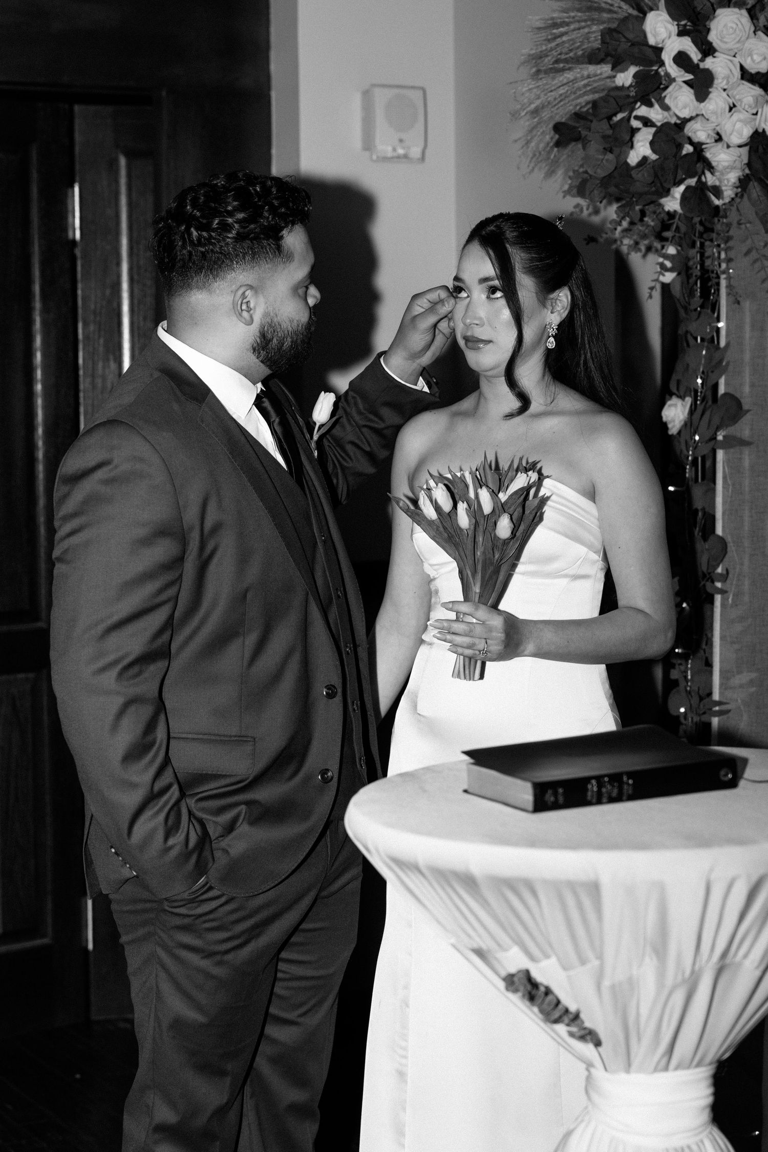 Groom gently wipes bride's tear during emotional wedding ceremony, bride holds tulip bouquet in elegant white gown.