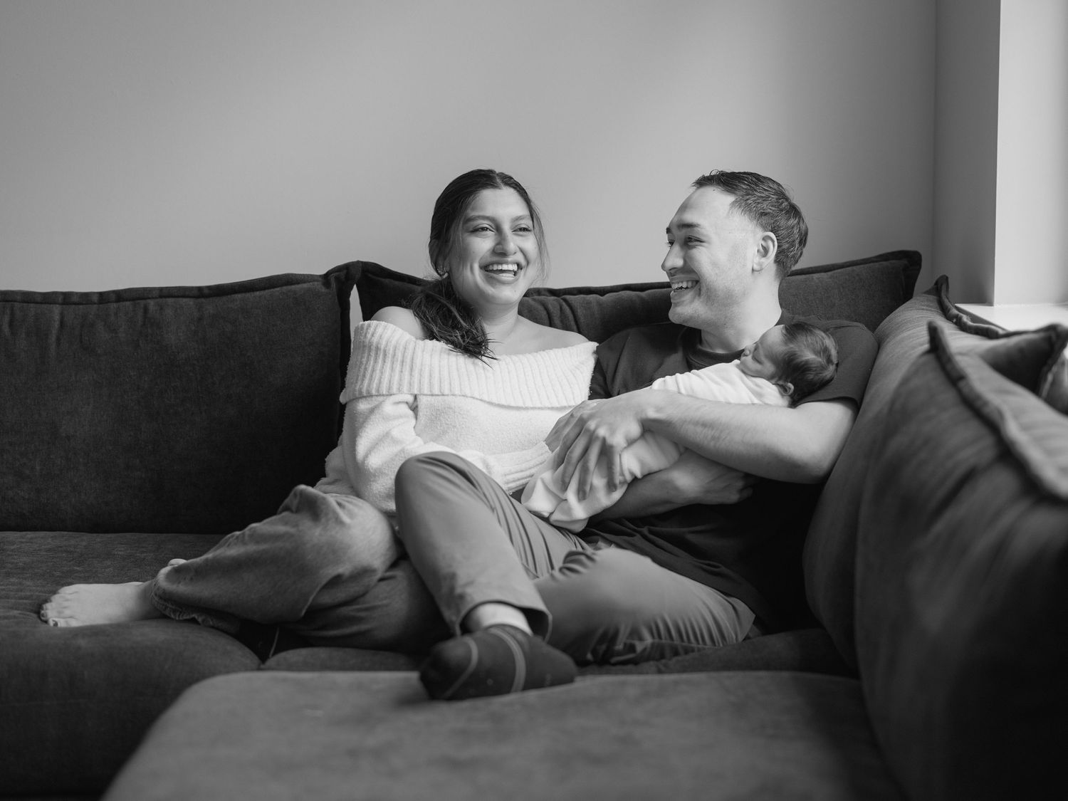 Happy couple sitting on a dark sofa, laughing together while holding a newborn baby in a cozy home.