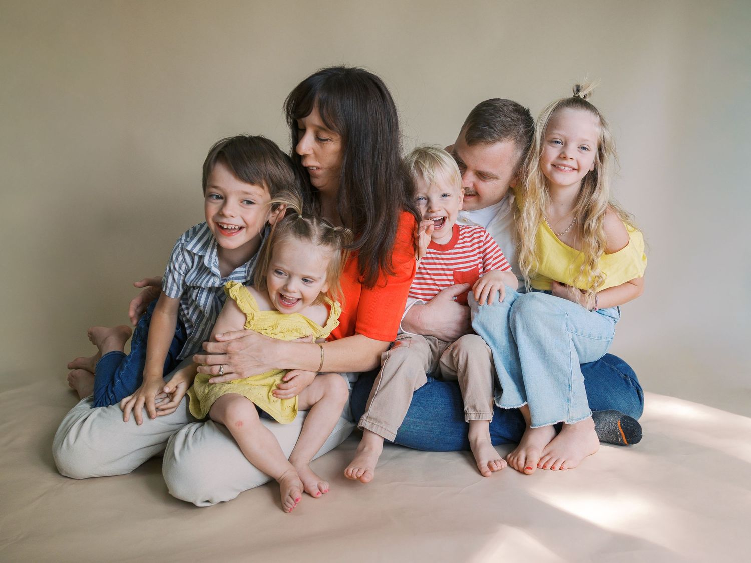 A family of six poses together on a studio floor, with two adults and four young children wearing colorful outfits.