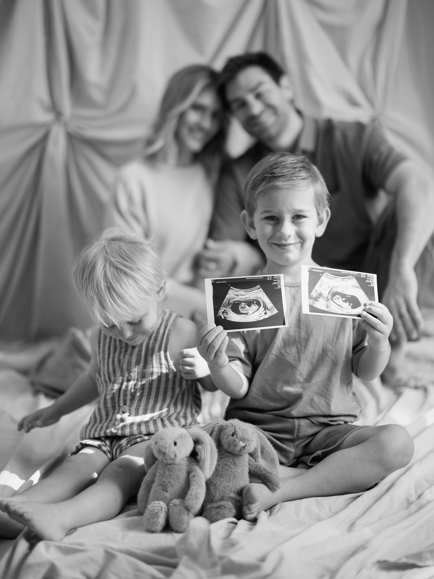 Black and white family photo with two boys holding ultrasound pictures, parents embracing in background on bed.