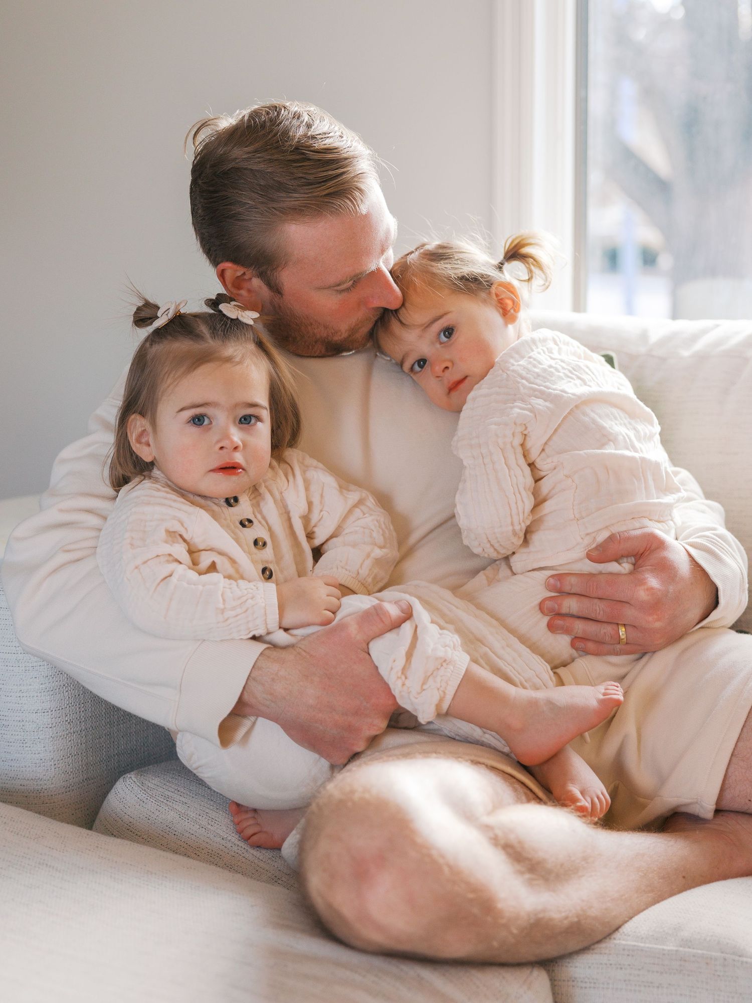 A bearded father sits on a couch hugging two toddler girls in matching cream outfits near a bright window.