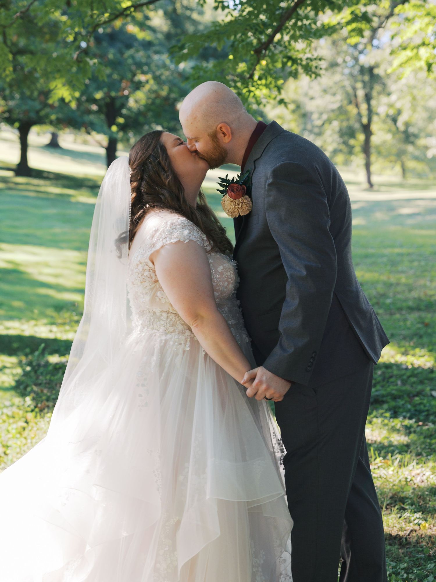 Bride and groom sharing a kiss outdoors on their wedding day, surrounded by lush green trees.