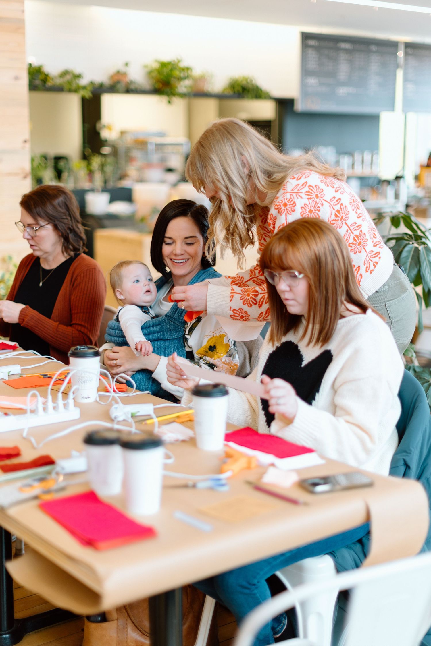 Women gather at a table for a crafting workshop, with one holding a baby, surrounded by supplies and coffee cups.