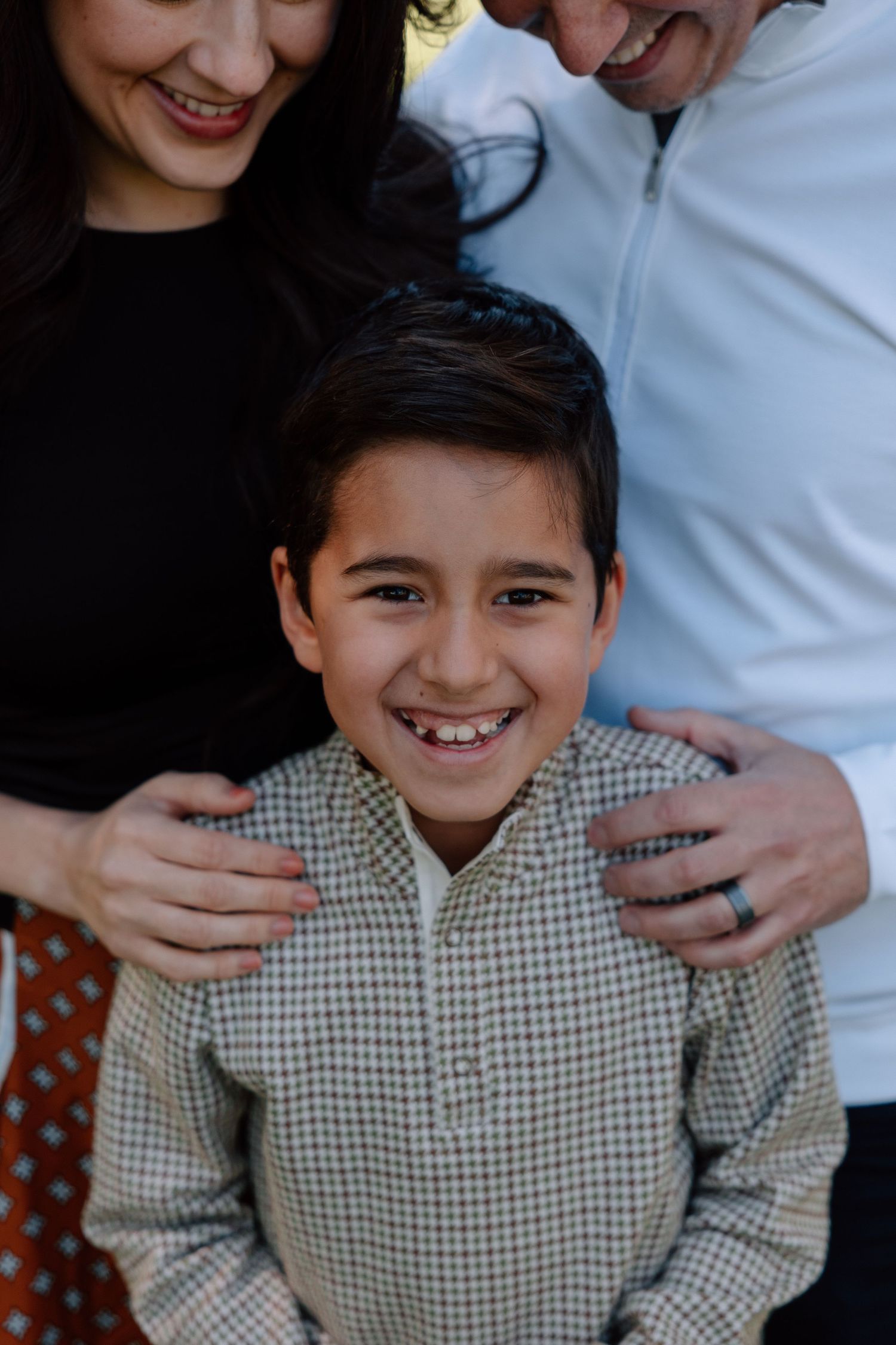 A smiling boy in a checkered shirt poses with two adults resting their hands on his shoulders in a family portrait.