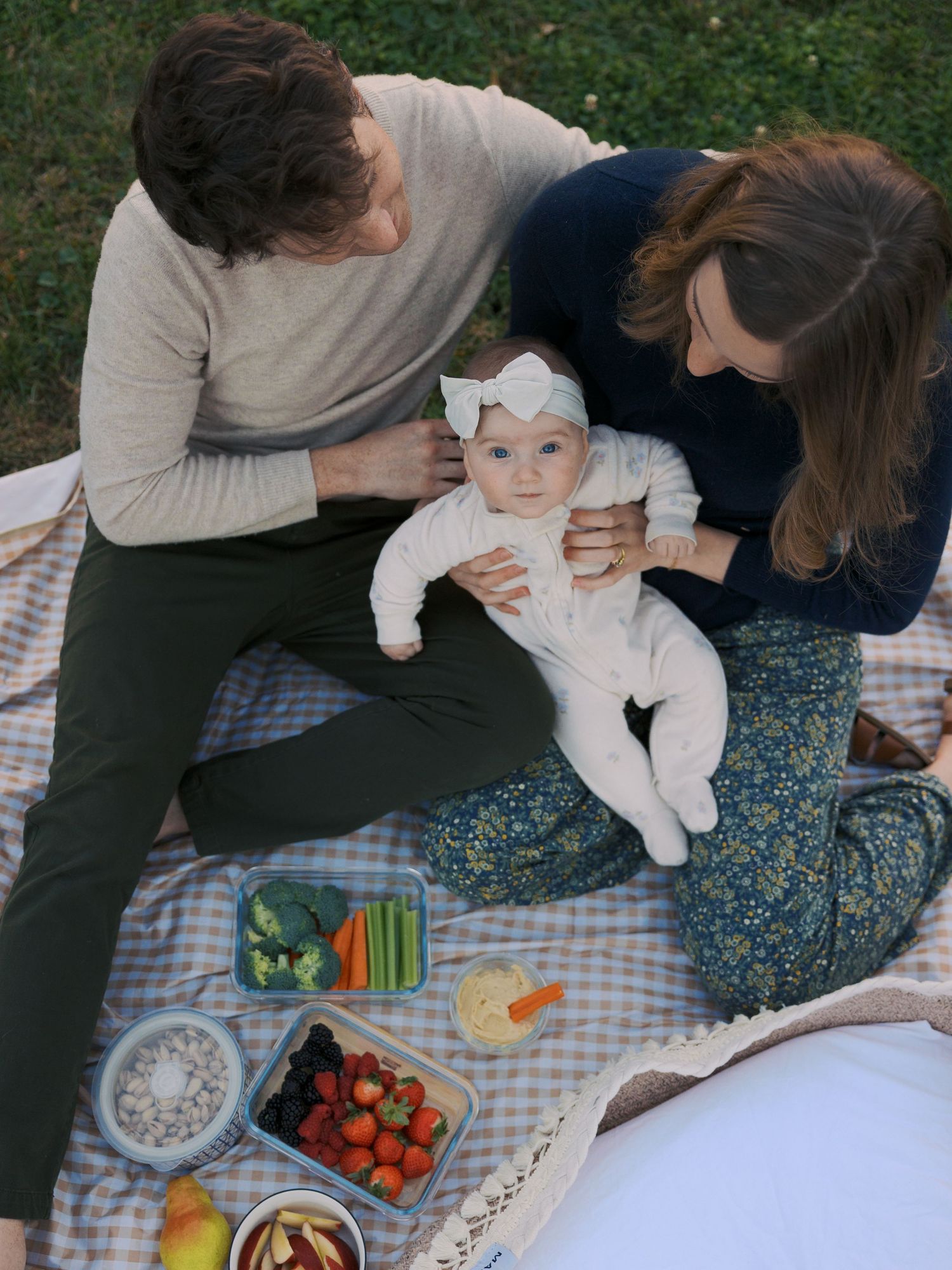 Family having an outdoor picnic on a checkered blanket with fresh vegetables and snacks.