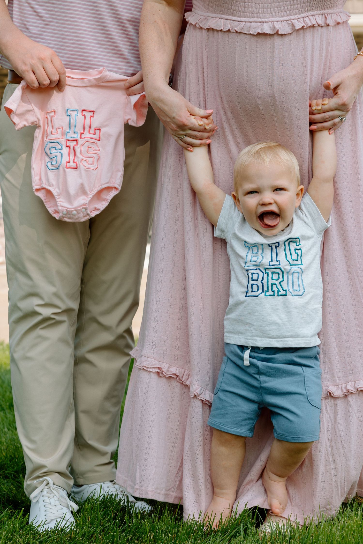 Toddler boy in Big Bro shirt stands with pregnant mom holding Lil Sis onesie, gender reveal photo.