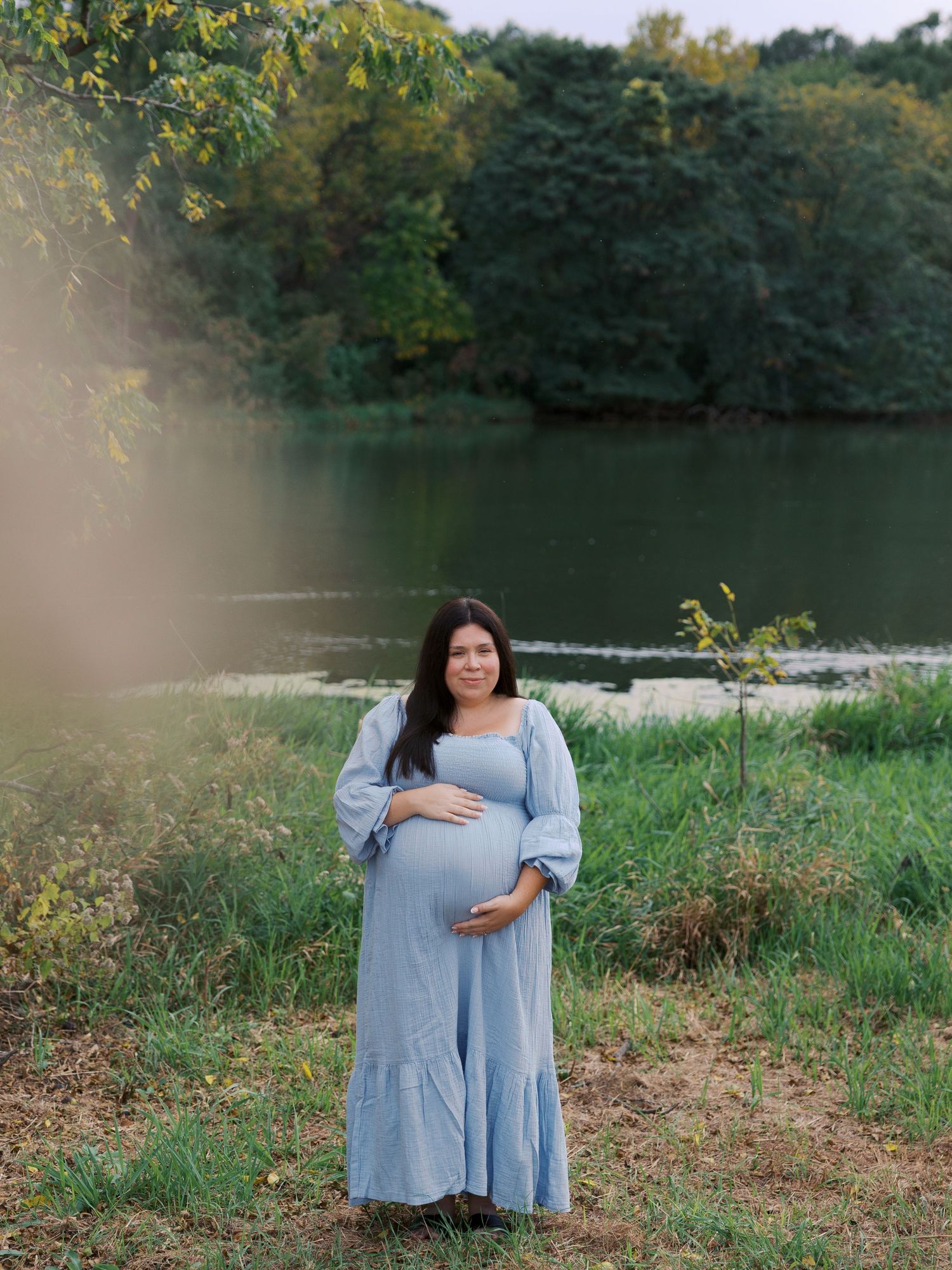 Pregnant woman in blue dress cradling baby bump by a serene lake surrounded by lush green trees.