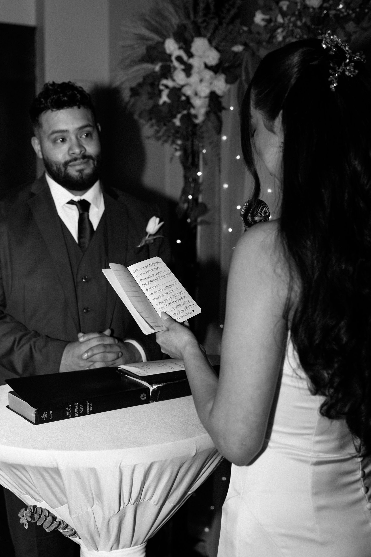 Black and white wedding photo of groom watching bride read vows from paper at elegant ceremony.