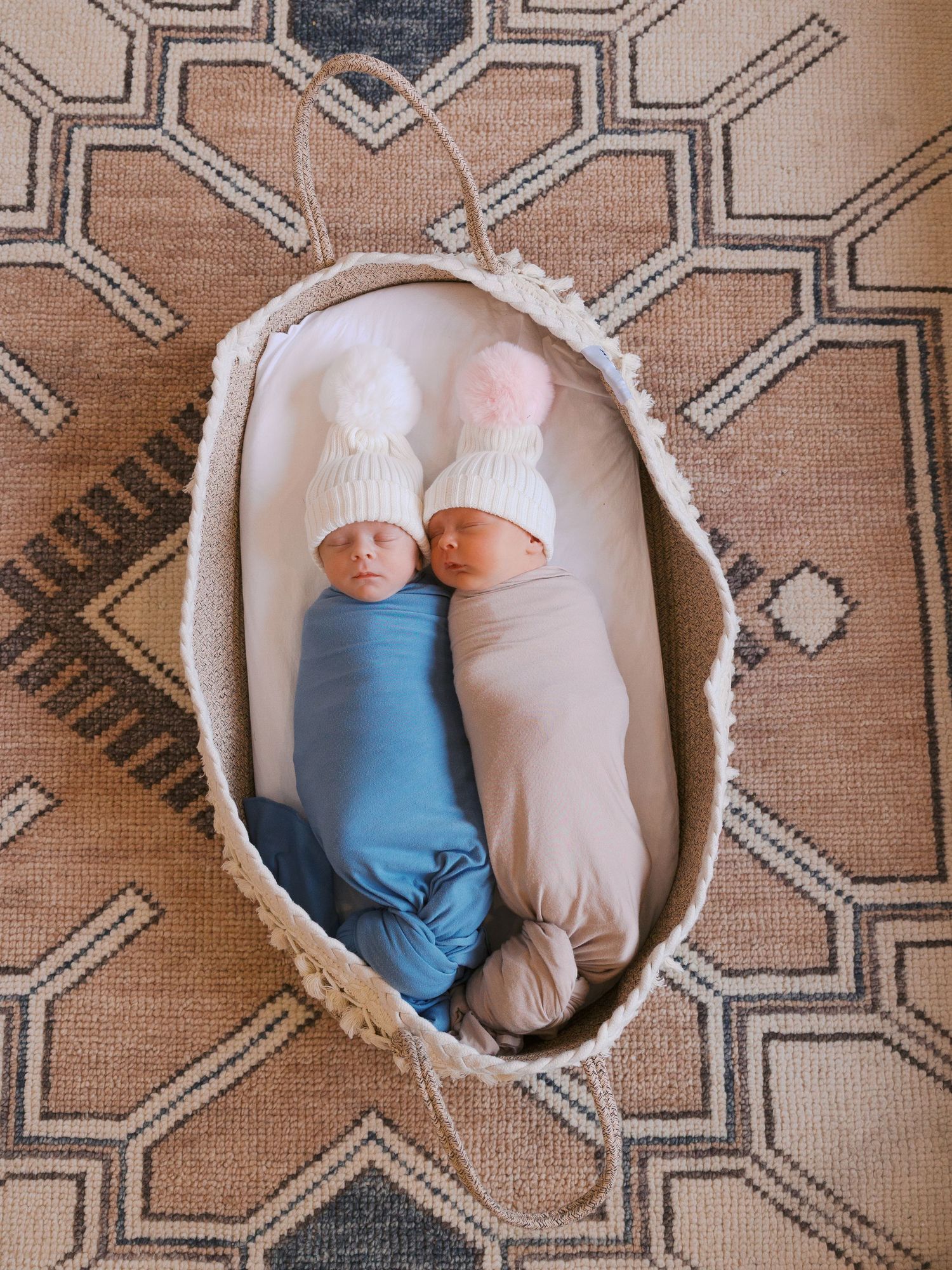 Two swaddled newborn twins wearing white pom-pom hats rest in a woven Moses basket on a patterned rug.