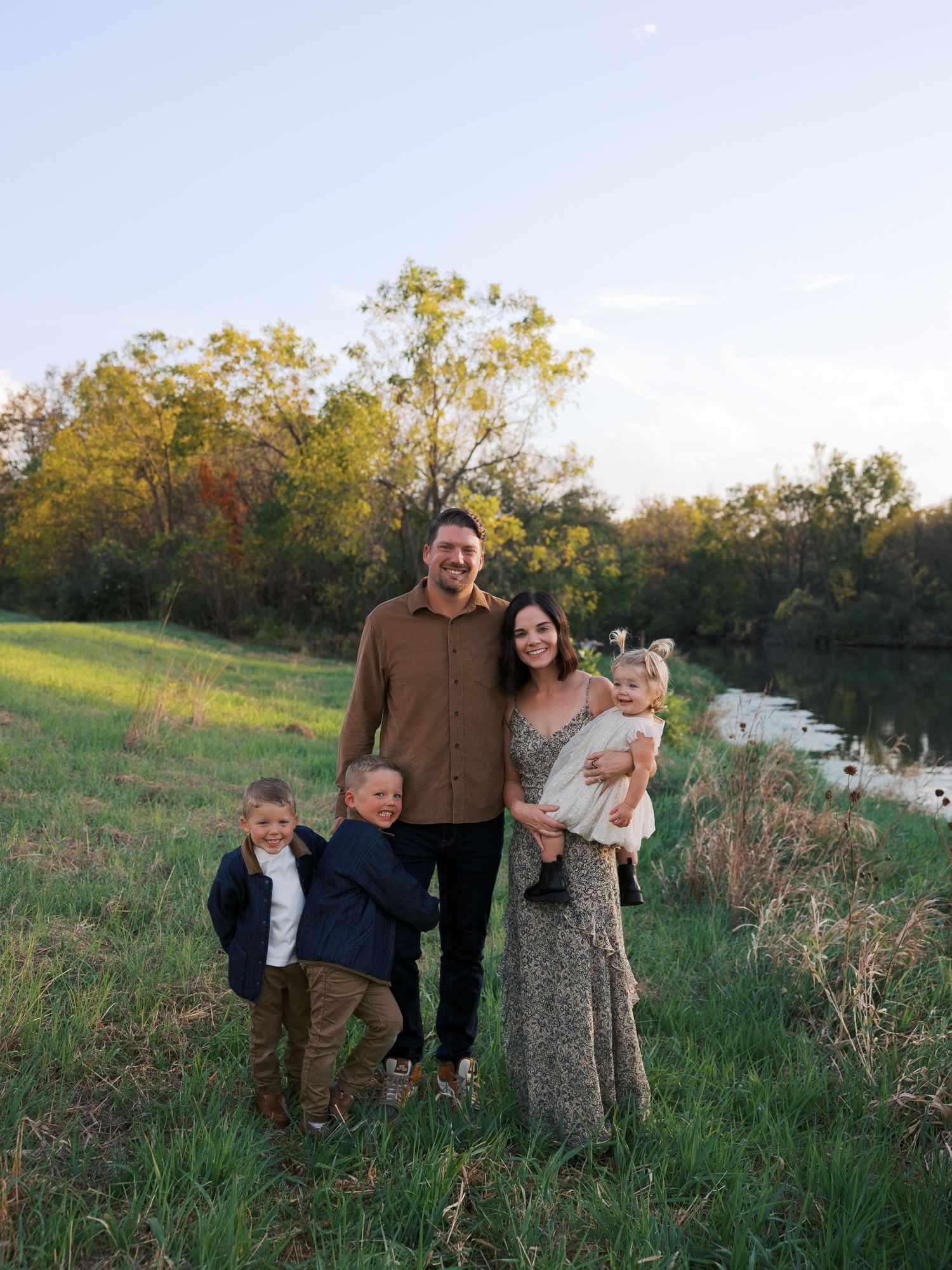 Family of five posing outdoors in a green meadow near a pond, surrounded by autumn trees at dusk.