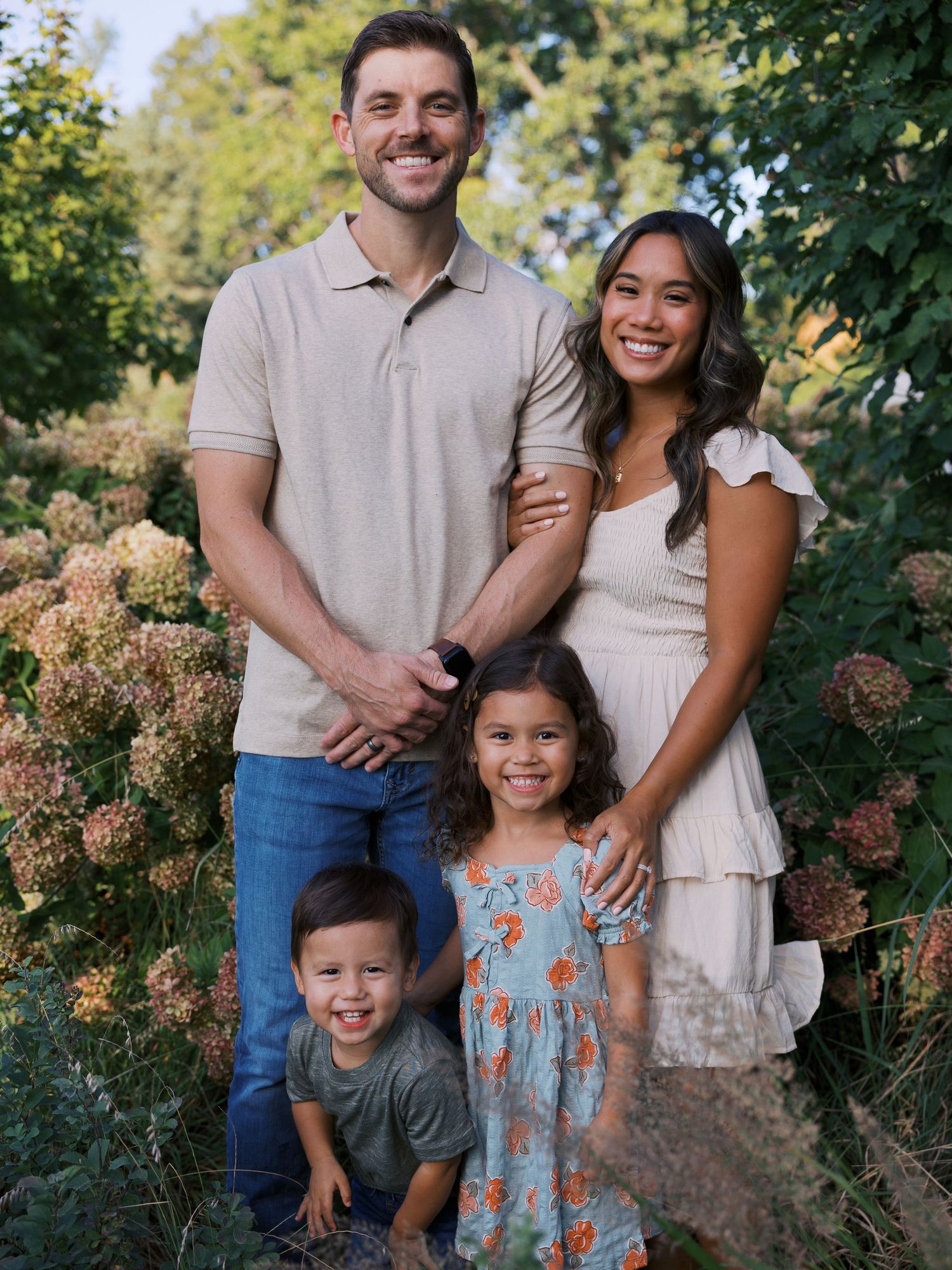 A smiling family of four poses outdoors among lush greenery and hydrangeas, with two young children in front.